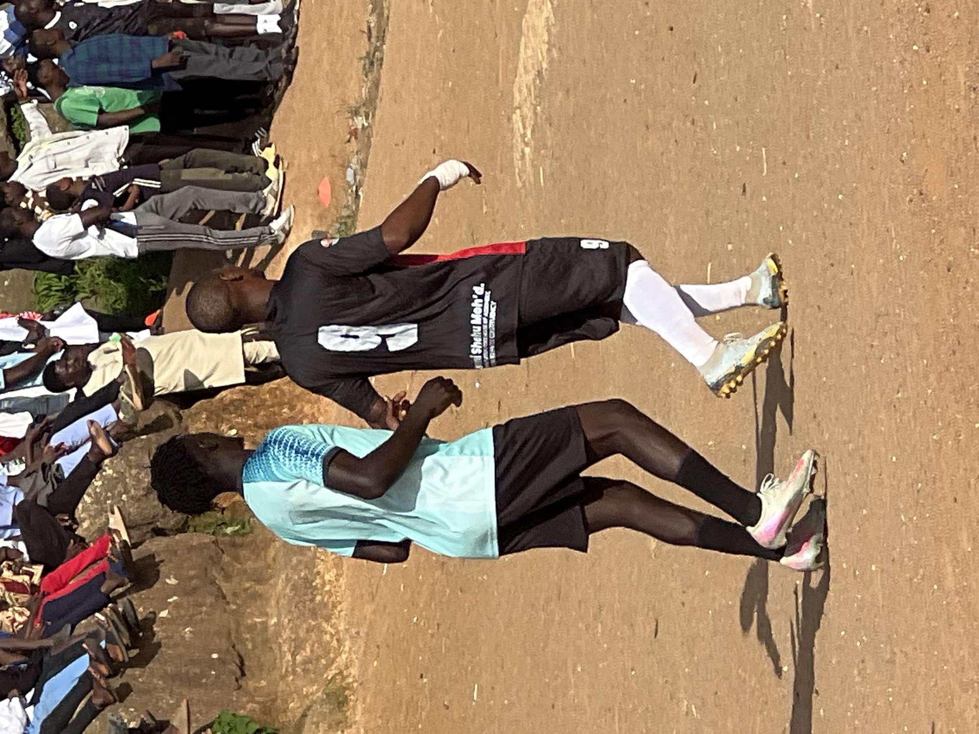 This image captures a street soccer match or practice in Jos, Nigeria. Two young men, wearing soccer attire, are the central focus. The man on the left wears a light blue and white jersey with black shorts and pink and white soccer cleats. The man on the right sports a black jersey with red accents, black shorts, white socks, and multi-colored soccer cleats. His jersey has the number "9" prominently displayed on the back, along with text that appears to be a name and possibly a sponsorship.
In the background, a group of spectators is seated on a rocky, elevated area, observing the activity. They are dressed in various casual clothing, reflecting a common scene at informal sporting events. The ground is a reddish-brown dirt, typical of many playing fields in the region. The overall environment suggests an outdoor, community-based setting.