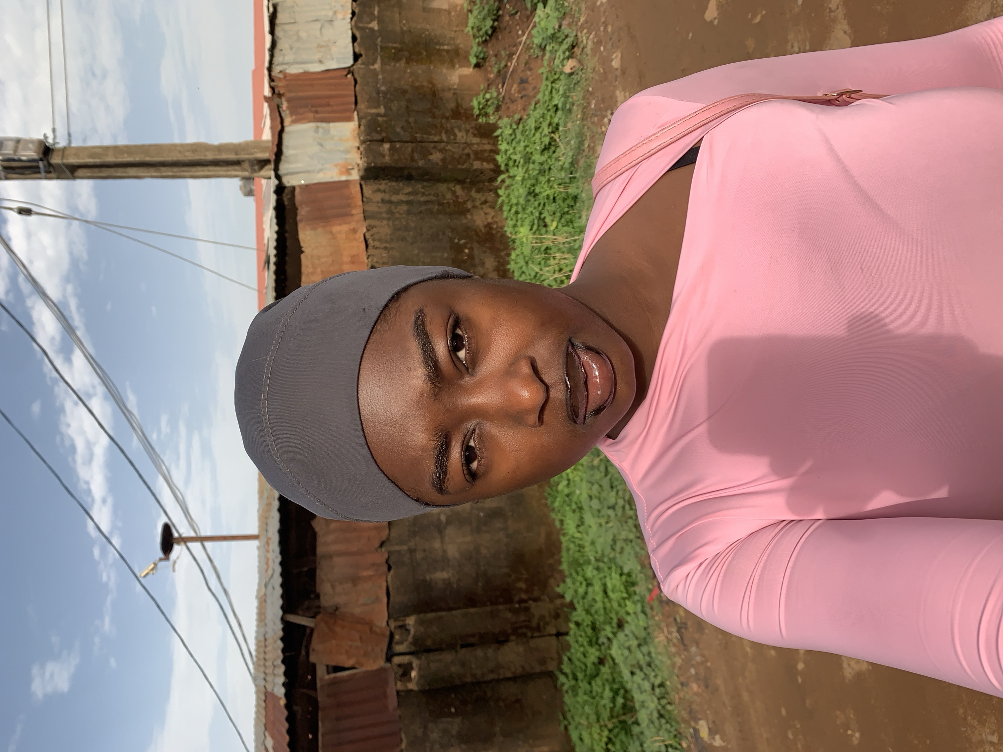 A person wearing a pink top and gray head covering stands outdoors. A power pole with wires is visible on the left, along with corrugated metal structures and some green vegetation. The location is Rafin Par, Nigeria.