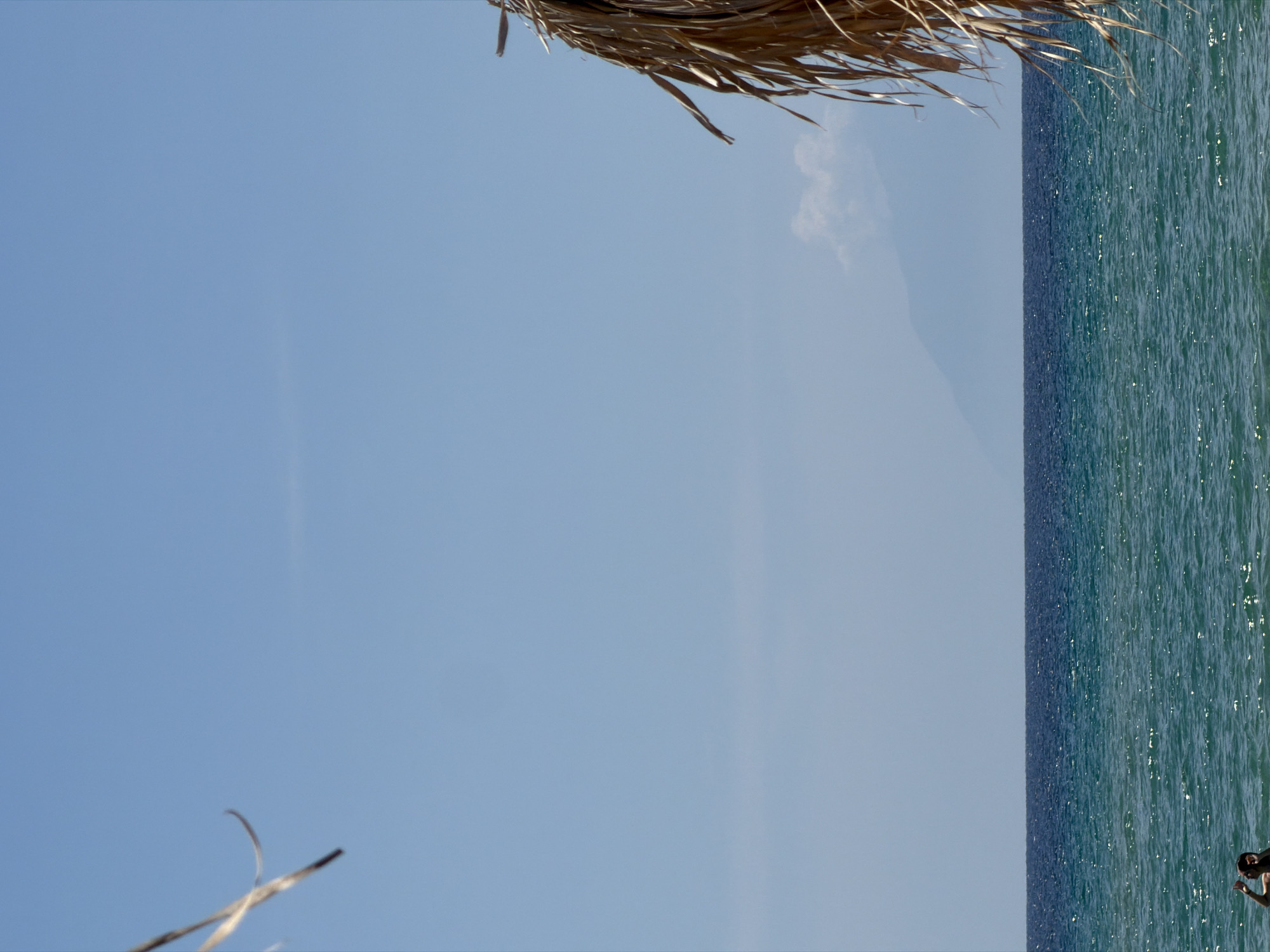 This image shows a view of the sea and sky in Nea Peramos, Greece, taken from under a straw sun umbrella. The sky is light blue with a few faint clouds. The sea is a darker blue-green, with sunlight reflecting on the water's surface. A person can be seen in the bottom right corner of the image. The edge of a straw sun umbrella is visible at the top of the image.