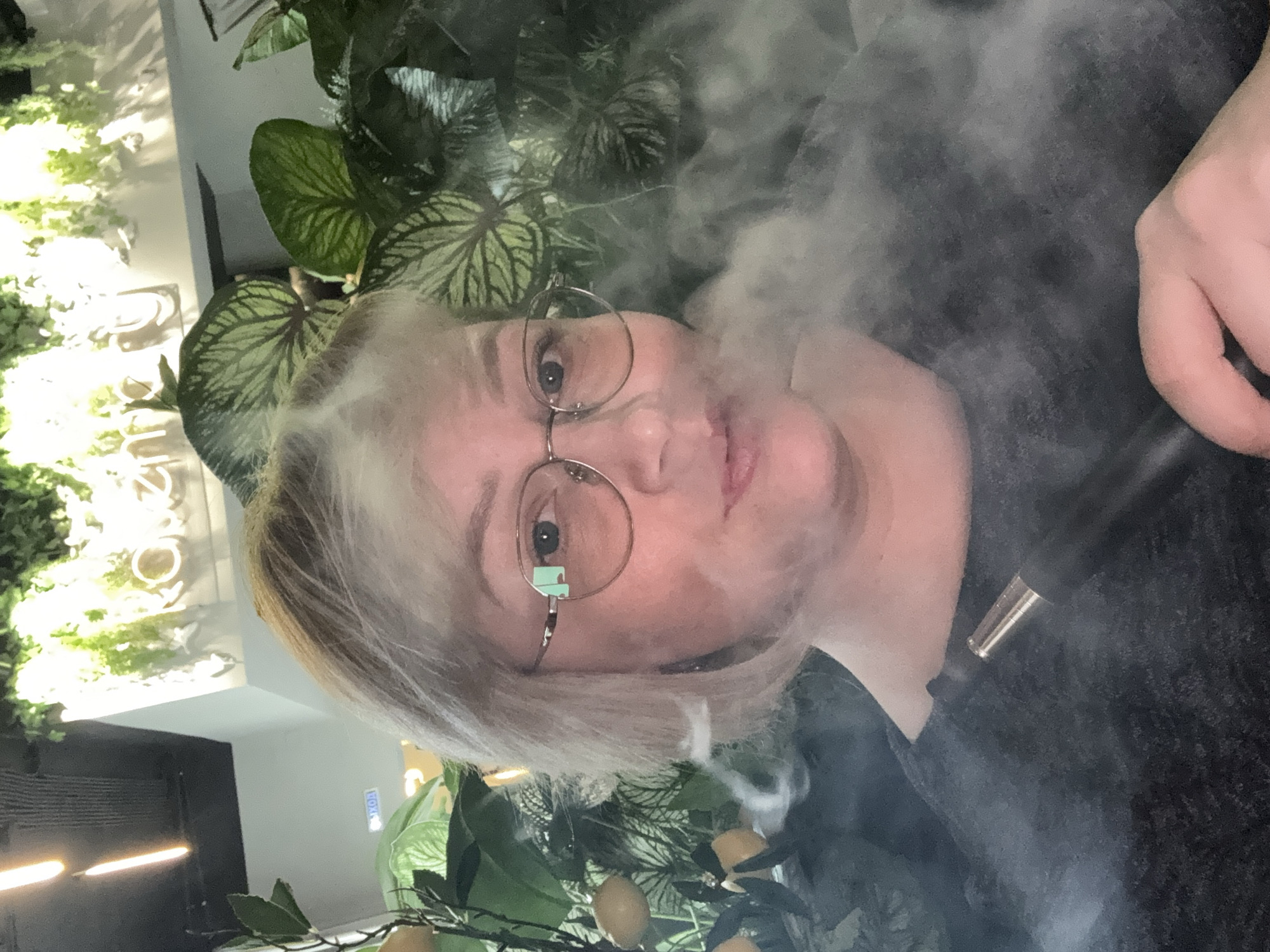 In this close-up, selfie-style photograph, a Caucasian woman with short, blonde hair and glasses is exhaling a cloud of vapor from a vaping device. She is wearing a dark, possibly grey, top. The background is a lush indoor environment with a variety of green plants, some with large leaves, and artificial lemons on a stem. A sign with glowing letters that appears to spell "roza" or a similar word is visible in the upper background, along with some lighting fixtures. There is also a blue sign with Russian text that reads "ВЫХОД" (VYKHOD), meaning "EXIT." The overall lighting suggests an indoor setting, possibly a cafe or lounge. There is no indication of Votkinsk, Russia, in the image itself.