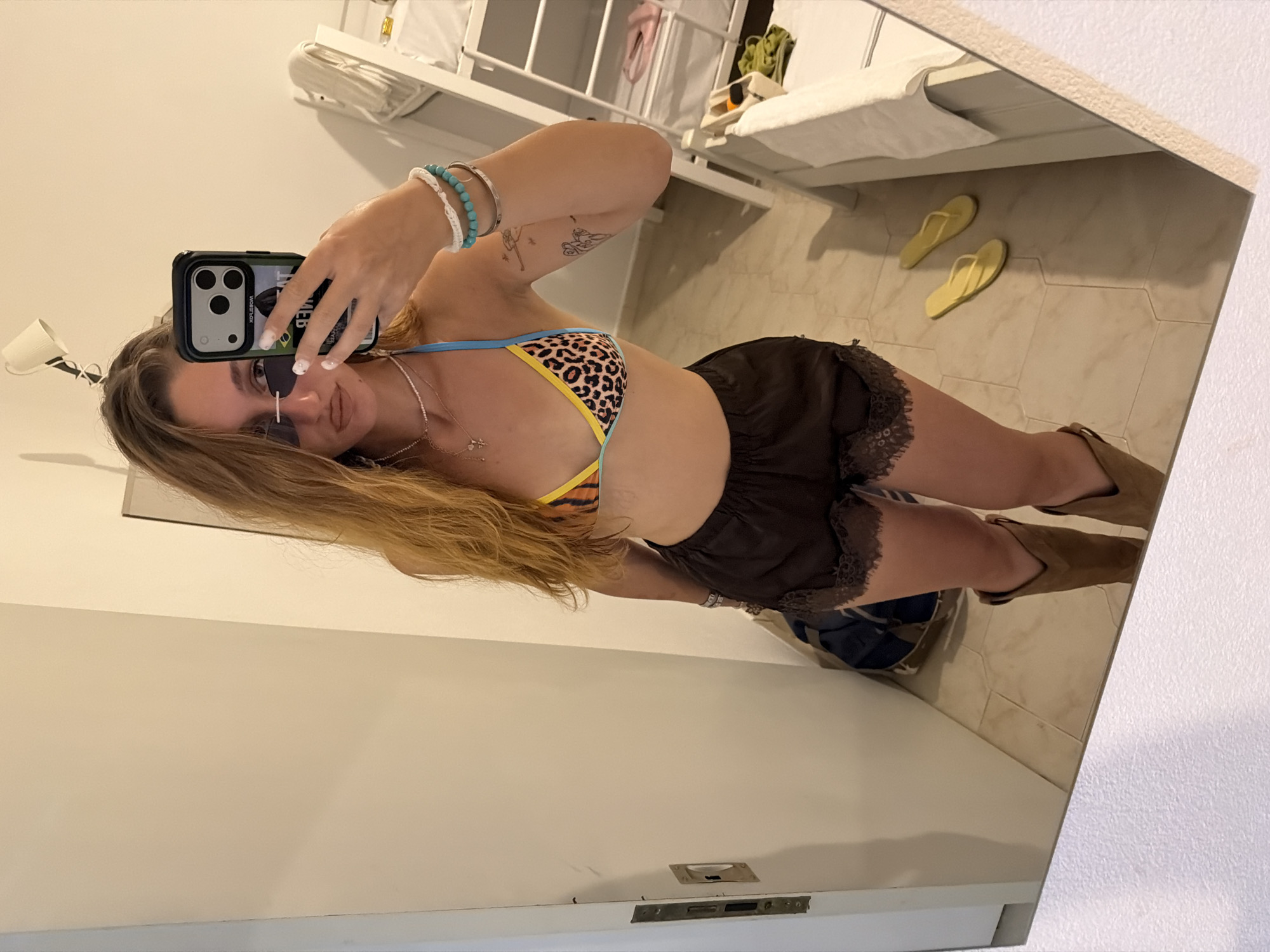 A young woman is taking a mirror selfie in what appears to be a hotel room. She is wearing a leopard print bikini top, dark shorts with lace trim, and brown cowboy boots. She has on sunglasses and several bracelets. In the background, a bunk bed is visible, along with a white towel and a pair of yellow flip-flops on the floor. The location is a hotel room in Stavros, Greece.