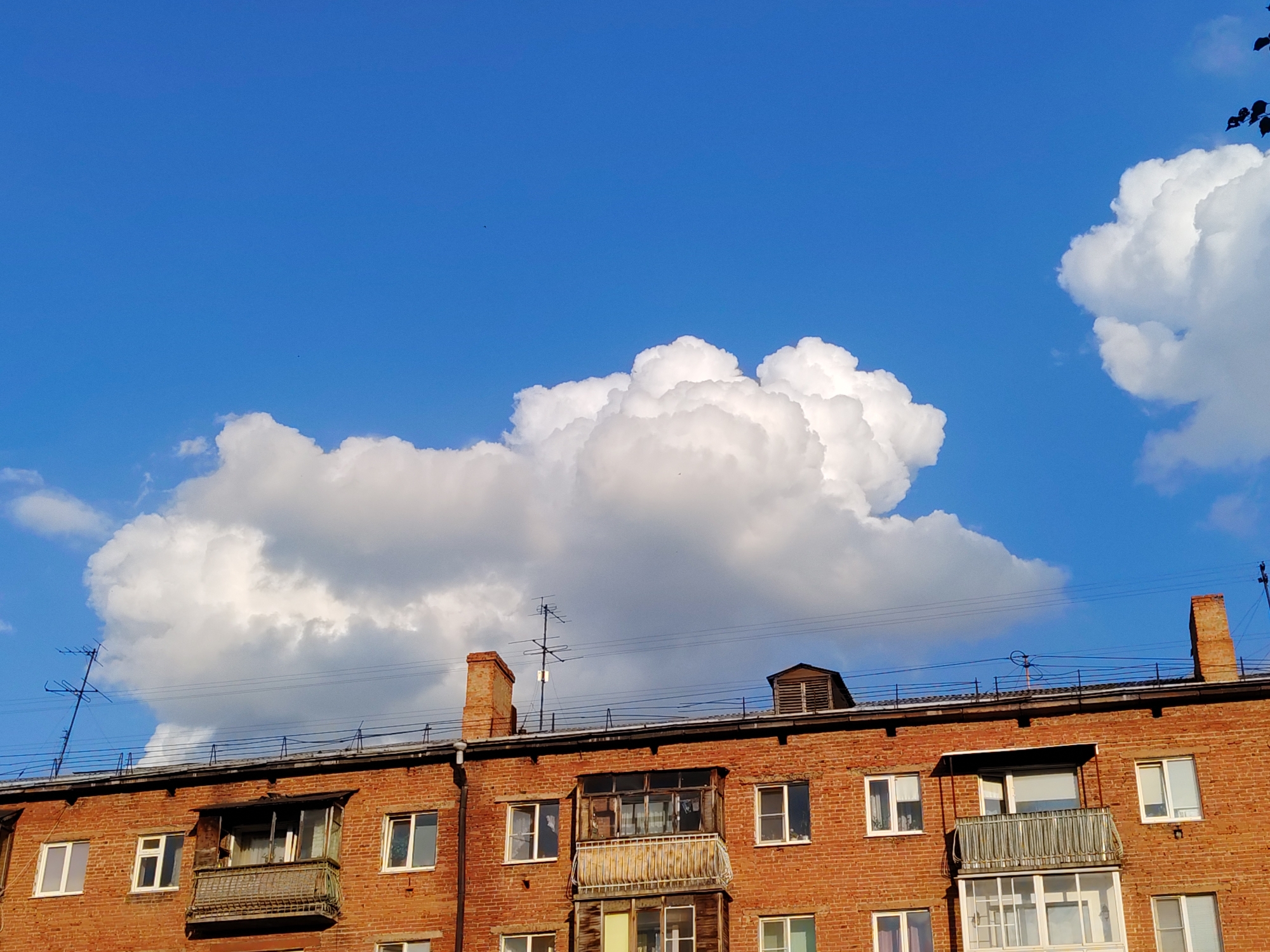 The image shows the upper floors of a red brick apartment building against a bright blue sky filled with large, fluffy white clouds. The building has several windows and balconies. On the roof, there are chimneys, antennas, and what appears to be a small attic structure. The scene is likely located in Kemerovo, Russia, given the architectural style of the building and the presence of a TV antenna, which is common in older residential areas. No people are visible in the frame.