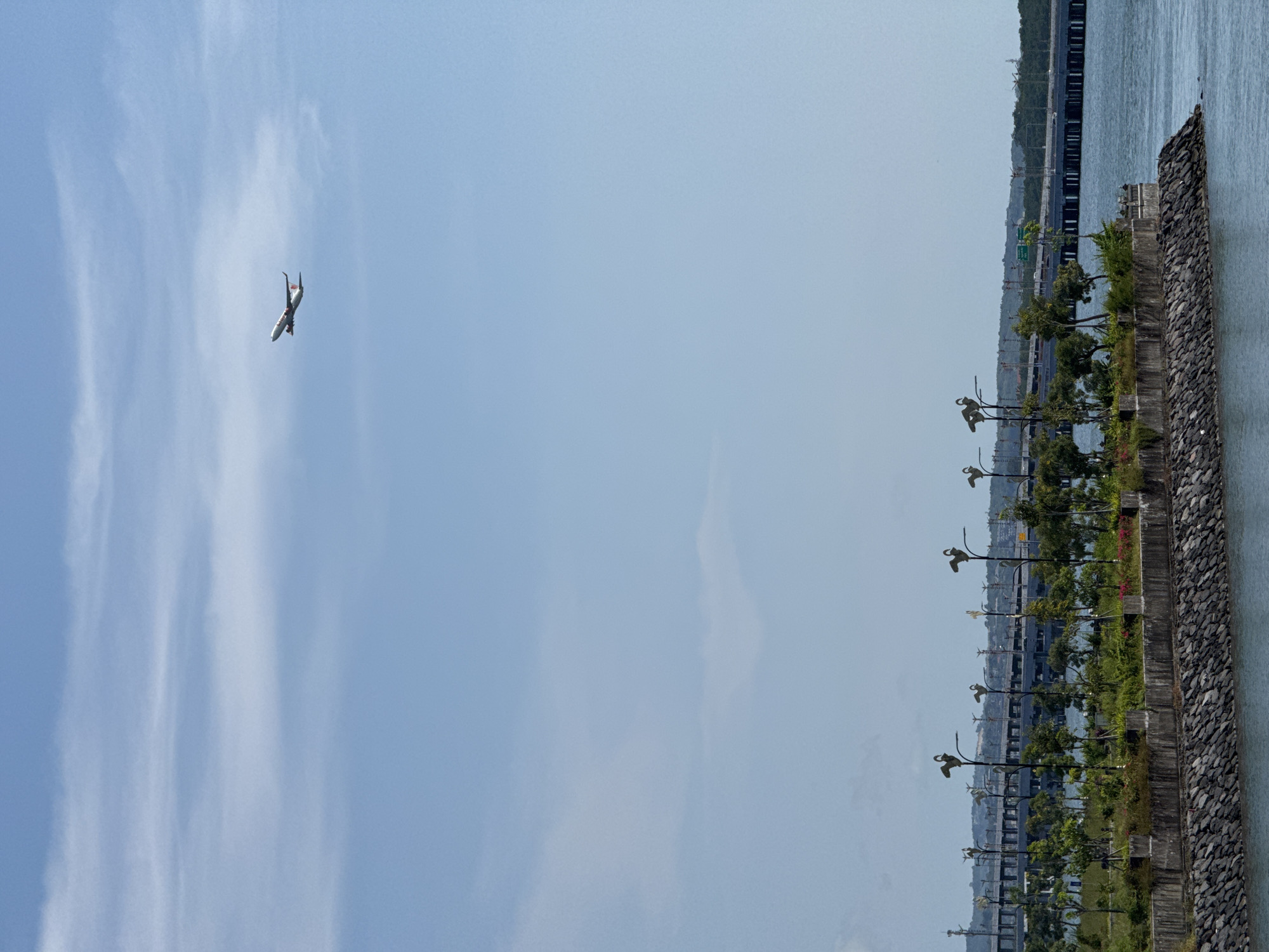 The image shows an airplane in the sky above the city of Denpasar, Indonesia. The sky is light blue with streaky clouds. The airplane is on the left of the image. On the right side of the image is a view of a strip of green grass and trees, a stone wall, and water. In the background are buildings and a bridge. There are lamp posts along the grass strip.