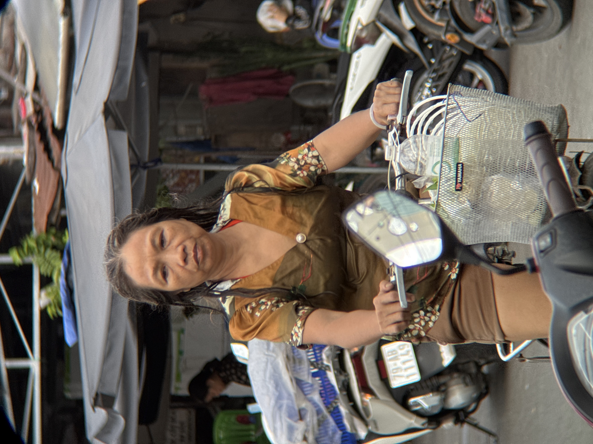 The image captures a woman riding a motorcycle in what appears to be an urban environment, possibly a market or street in Nha Trang, Vietnam. The woman has dark, wet hair and is wearing a tan and gold patterned blouse over a red undergarment, and brown shorts. She is holding the motorcycle's handlebars, with a basket attached to the front, containing a water bottle with a Yamaha logo. The motorcycle's license plate reads "79-AB 111.49". In the background, there are other motorcycles, various stalls or awnings providing shade, and some foliage. The overall atmosphere suggests a busy street scene.
