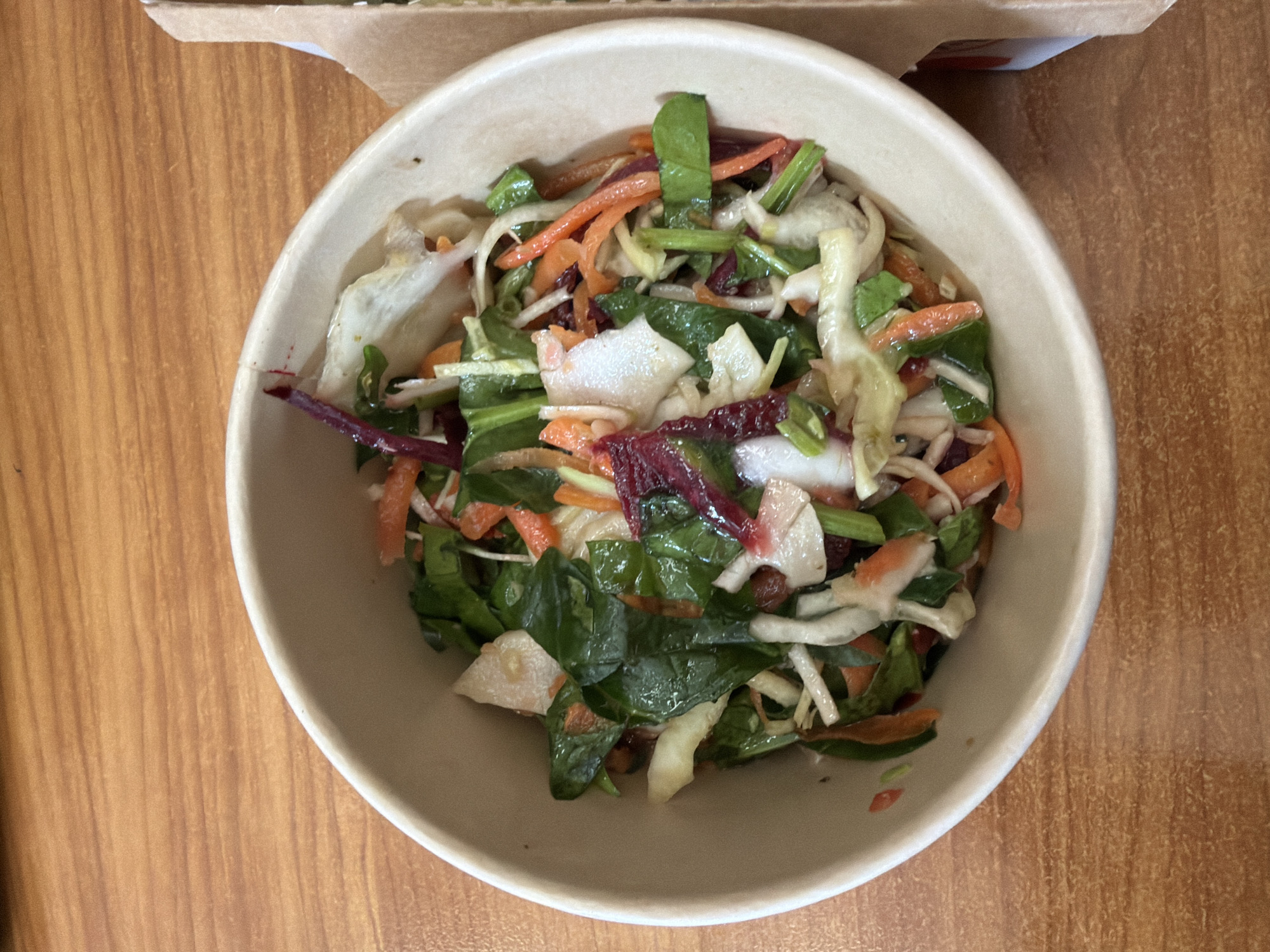 A top-down shot of a bowl of salad on a light brown wooden surface. The bowl is off-white and contains a salad consisting of shredded carrots, dark green leafy vegetables (possibly spinach), shredded cabbage, sliced beets, and pieces of what appears to be a white root vegetable or possibly pear or apple. A portion of a cardboard takeout container can be seen in the background. There is no evidence to suggest that this image was taken in Sofia, Bulgaria.