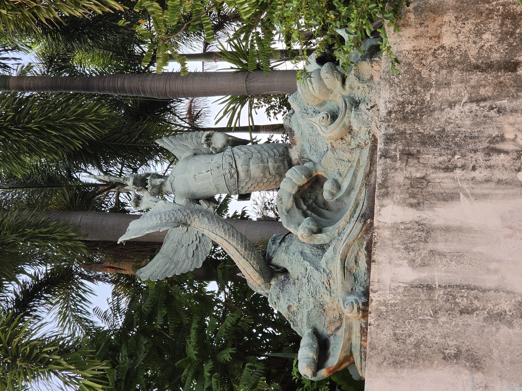 The image shows a statue of a winged creature with a fish-like tail, situated next to a weathered, light-colored wall. The statue has detailed carvings, including scales on the tail and feathers on the wings. Palm trees and other greenery are visible in the background. The location is Pattaya City, Thailand.