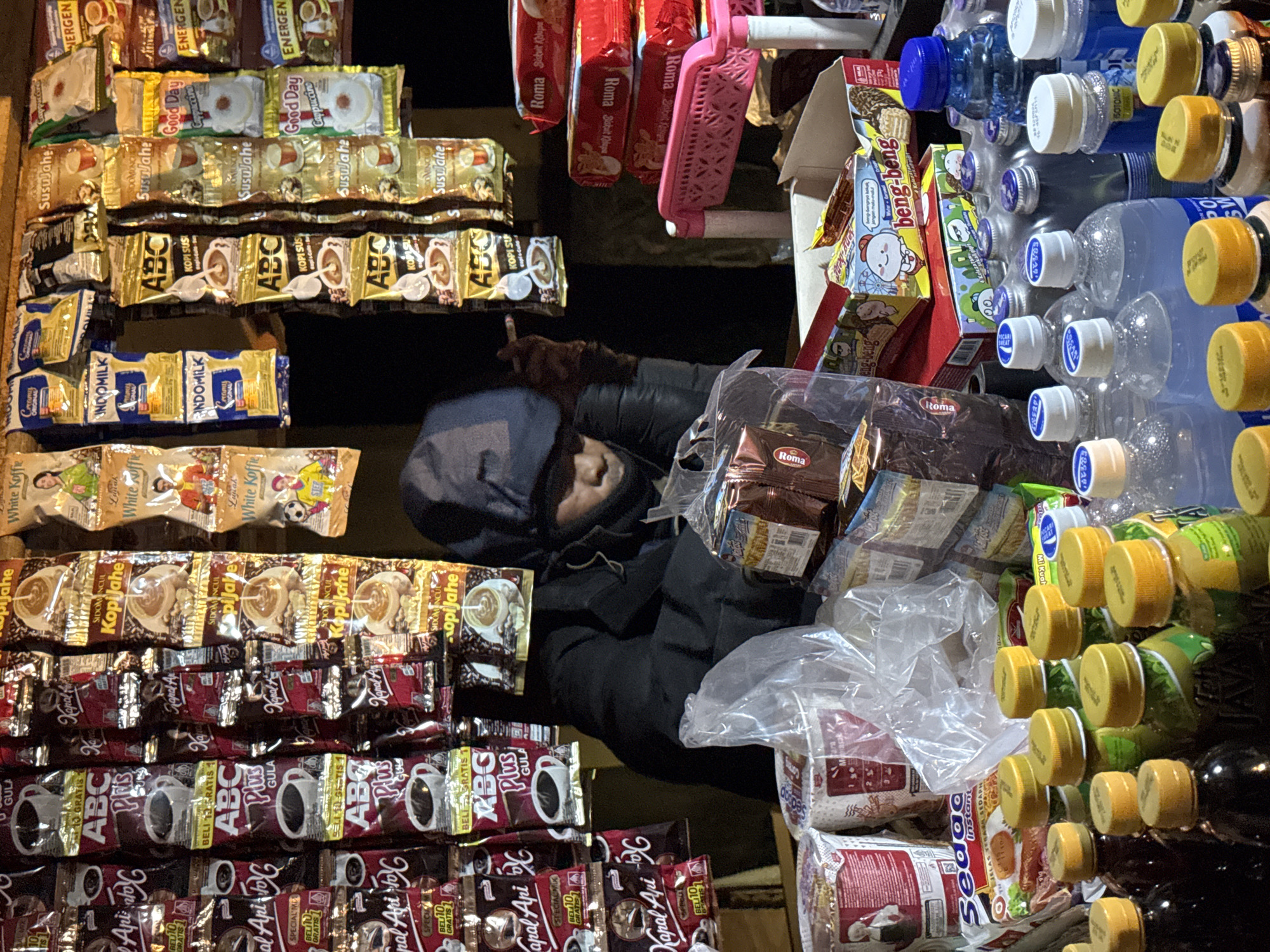 The image shows a street vendor in Unknown, Indonesia, surrounded by a variety of products. A person wearing a dark hooded jacket is visible behind the merchandise, possibly the vendor. The foreground is filled with rows of stacked instant coffee and tea packets from brands like "ABC," "Kapal Api," "Indomilk," "Energen," "Good Day Cappuccino," and "White Koffie." To the right, there are stacks of bottled water with blue and yellow caps, along with bottles of a yellow drink and dark liquid. Also, there are boxes of "beng-beng" wafers, "Roma" biscuits, and packs of "Sedaa" instant noodles. A pink plastic organizer hangs among the products. The overall impression is of a small, cluttered roadside stall offering a range of convenience items.