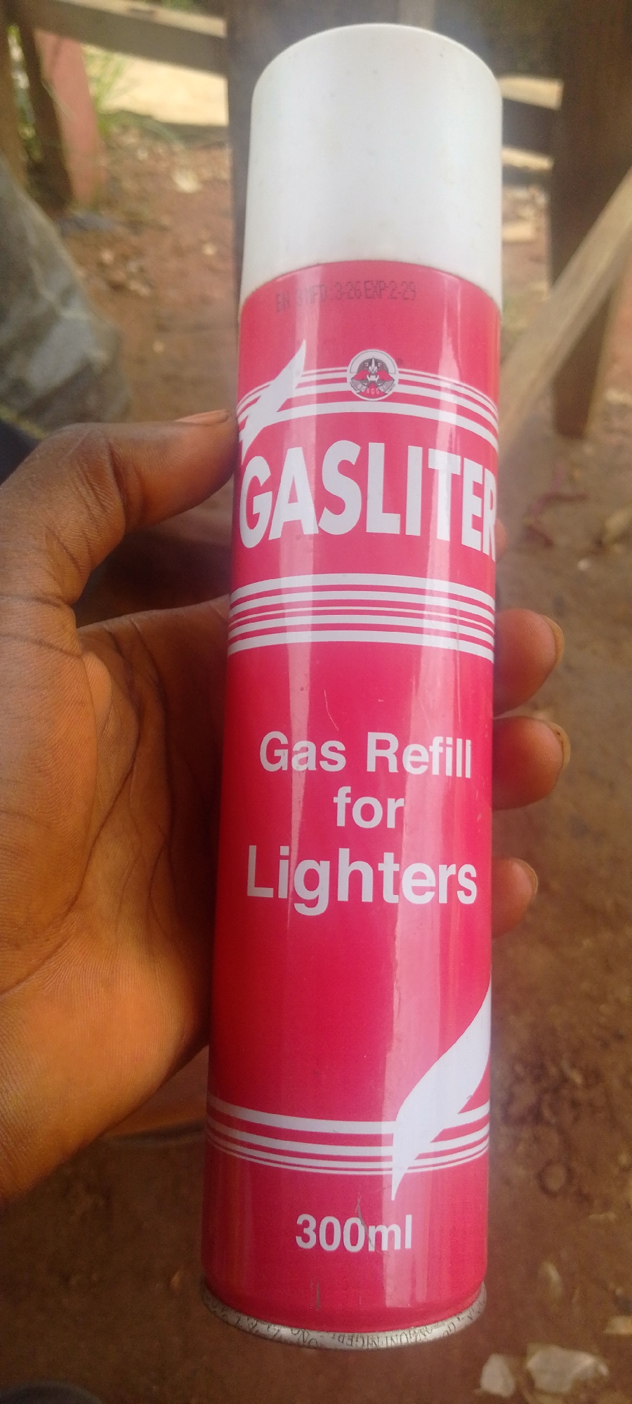 A close-up, vertical shot shows a hand holding a red and white can labeled "GASLITER" and "Gas Refill for Lighters" in English. The can also indicates "300ml" and has a small logo with a badger-like animal. There is text on the can that appears to be manufacturing and expiry dates, "MFD: 3-26 EXP: 2-29". The background is blurred and suggests an outdoor setting with earthy tones and some greenery, consistent with the environment in Baissa, Nigeria. The hand is of a person with dark skin, holding the can upright. The focus is on the can and the hand.