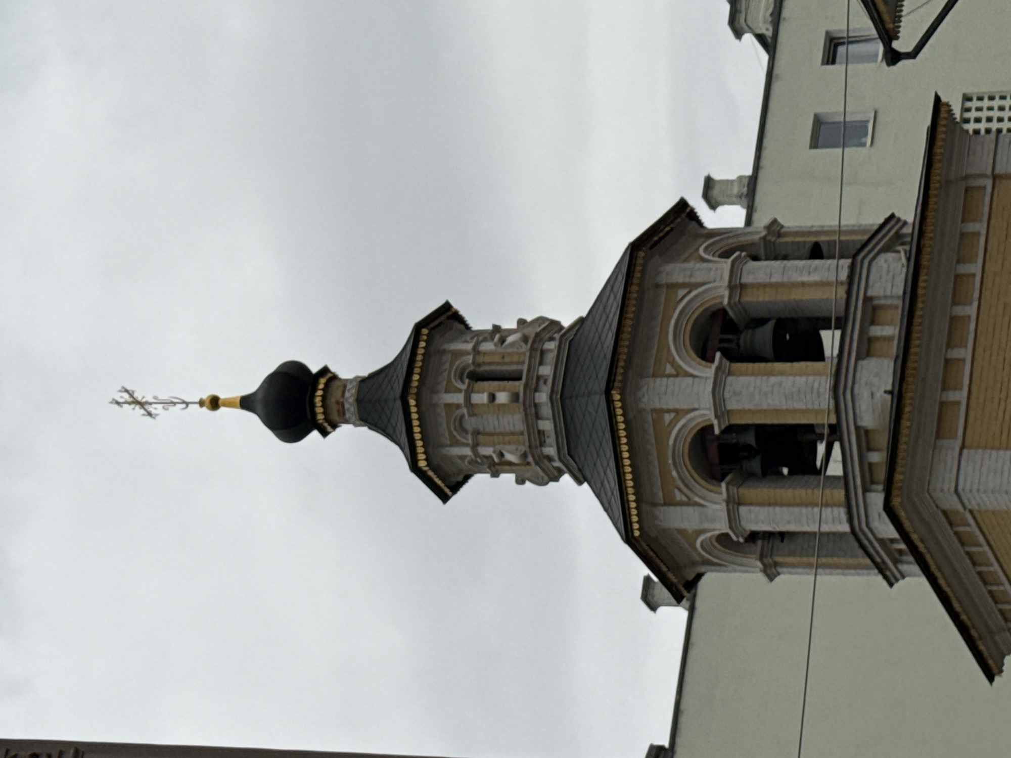 The image shows a close-up, low-angle view of a bell tower with a black, onion-shaped dome topped with a cross. The tower is multi-tiered and features yellow brickwork and white stone accents. Bells are visible through arched openings in the tower. In the background, parts of light-colored buildings with windows are visible under a cloudy sky. A thin horizontal wire cuts across the foreground, obscuring part of the bell tower. The scene is likely located in Moscow, Russia, given the architectural style of the bell tower, which is characteristic of Orthodox churches. There are no people visible in the image.