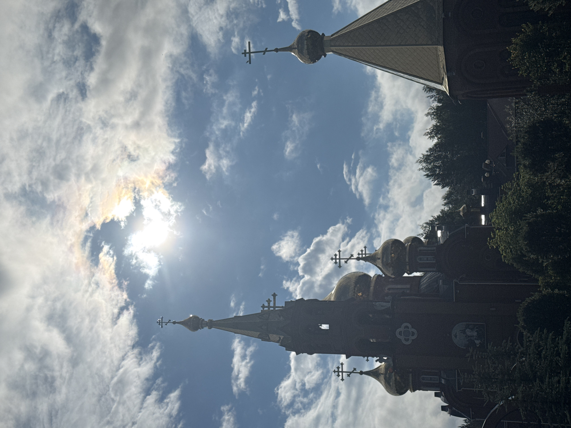 The image shows an exterior view of the Church of the Assumption of the Blessed Virgin Mary in Chaikovsky, Russia. The church features multiple onion domes, one of which is prominently gold-colored. The architecture is of a traditional Russian Orthodox style with red brick walls and ornate crosses topping the spires and domes. The sky is partly cloudy with the sun shining through, creating lens flare and highlighting the clouds. Trees surround the church, suggesting a location with natural greenery. No people are visible in the image.
