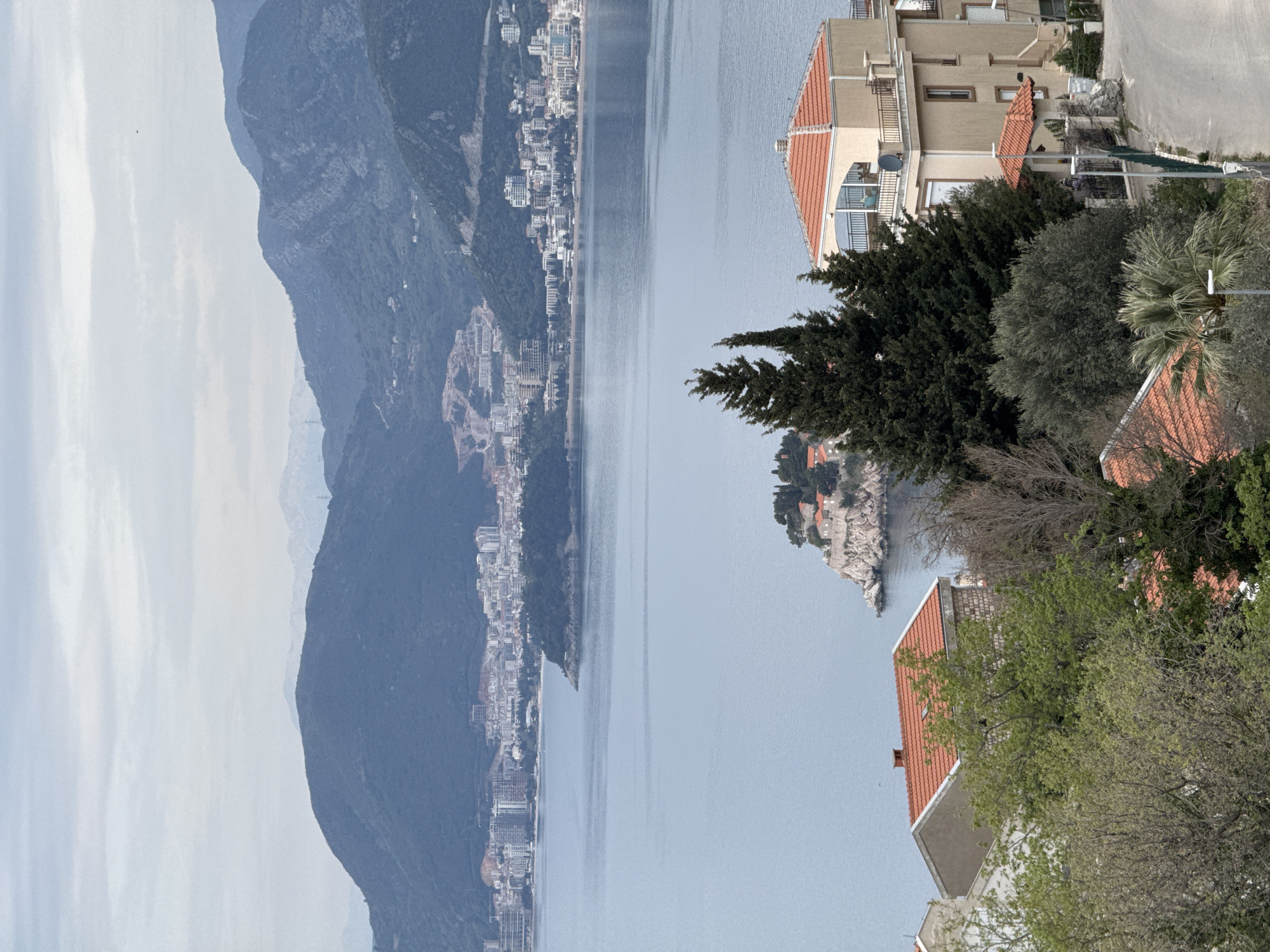 The image shows an elevated view in Blizikuće, Montenegro. In the background, there's a mountain range with a town built along its base and coastline. A large body of water stretches between the mountain and the foreground. Closer to the viewer, there are buildings with red tile roofs intermixed with trees and vegetation. The architecture is modern, with beige buildings, balconies and other modern design elements. An islet with vegetation and a few buildings stands in the water near the foreground.