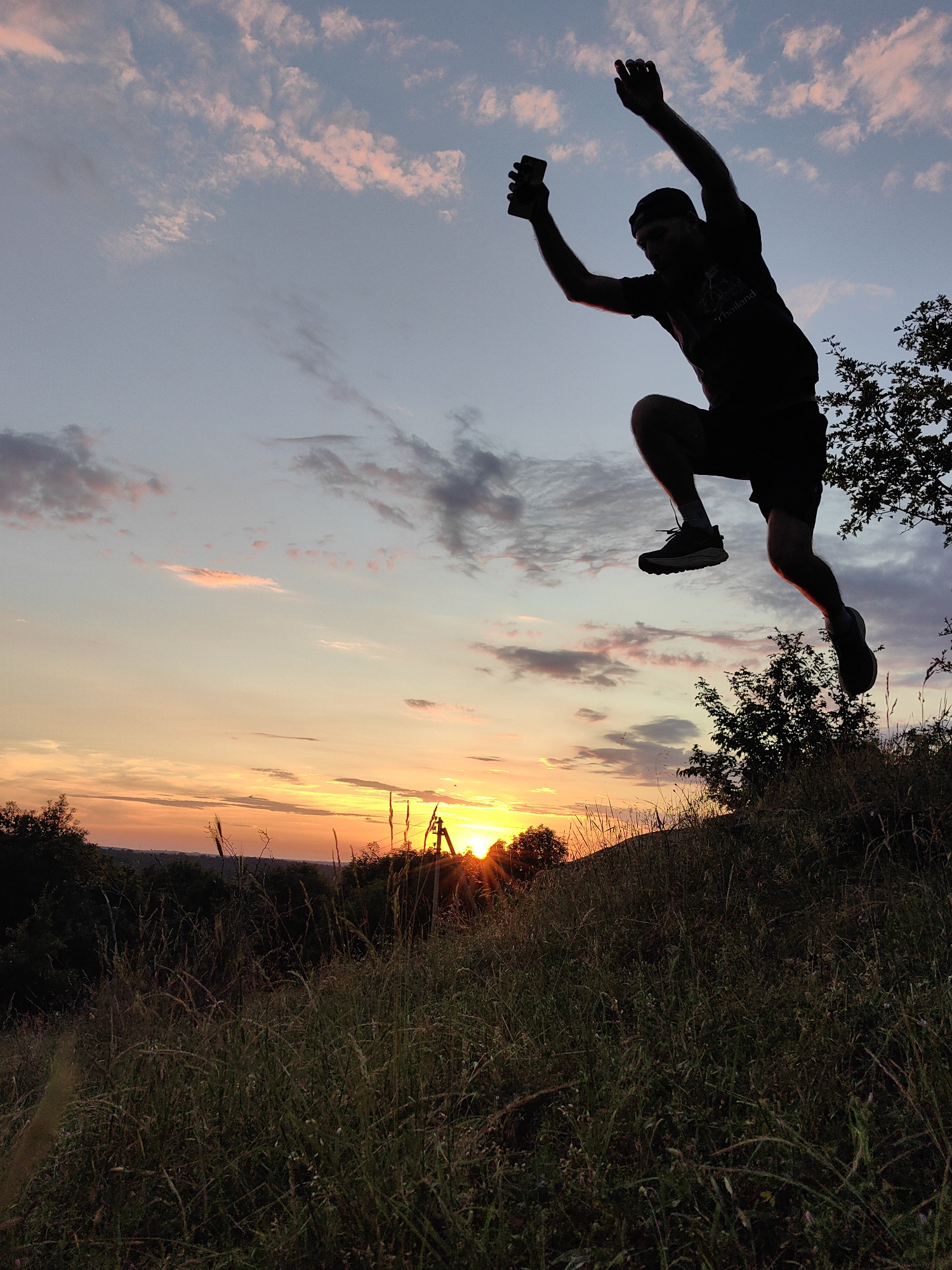 A silhouette of a person is captured mid-jump against a vibrant sunset sky in Зуйское сельское поселение, Ukraine. The sky is a gradient of soft blues, pinks, and oranges, with wispy clouds. The person, wearing shorts and a t-shirt, is holding a smartphone in their raised right hand. They are silhouetted against the bright sky, with their legs bent as if in the middle of an athletic leap. The foreground consists of a grassy hill, with tall, dry grass and some sparse trees on the right. The setting sun is visible on the horizon, casting a warm glow over the landscape.