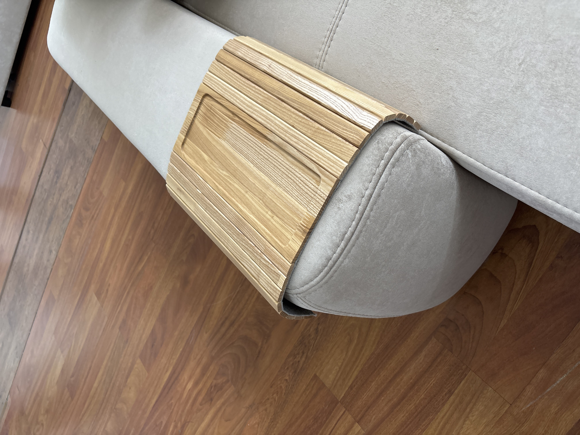 The image shows a close-up, eye-level, outdoor shot of a light gray sofa armrest with a wooden tray placed on top of it. The wooden tray has a slatted design with a rectangular indentation in the center. The sofa is positioned on a wooden floor with a warm, reddish-brown hue. The lighting is bright, suggesting it is daytime. The scene is devoid of any people, activity, or interaction. The background is slightly blurred, but appears to be an indoor setting with more wooden flooring visible. There is no indication of the city of Zaporizhzhia, Ukraine, or any elements that would place the image in that specific location.