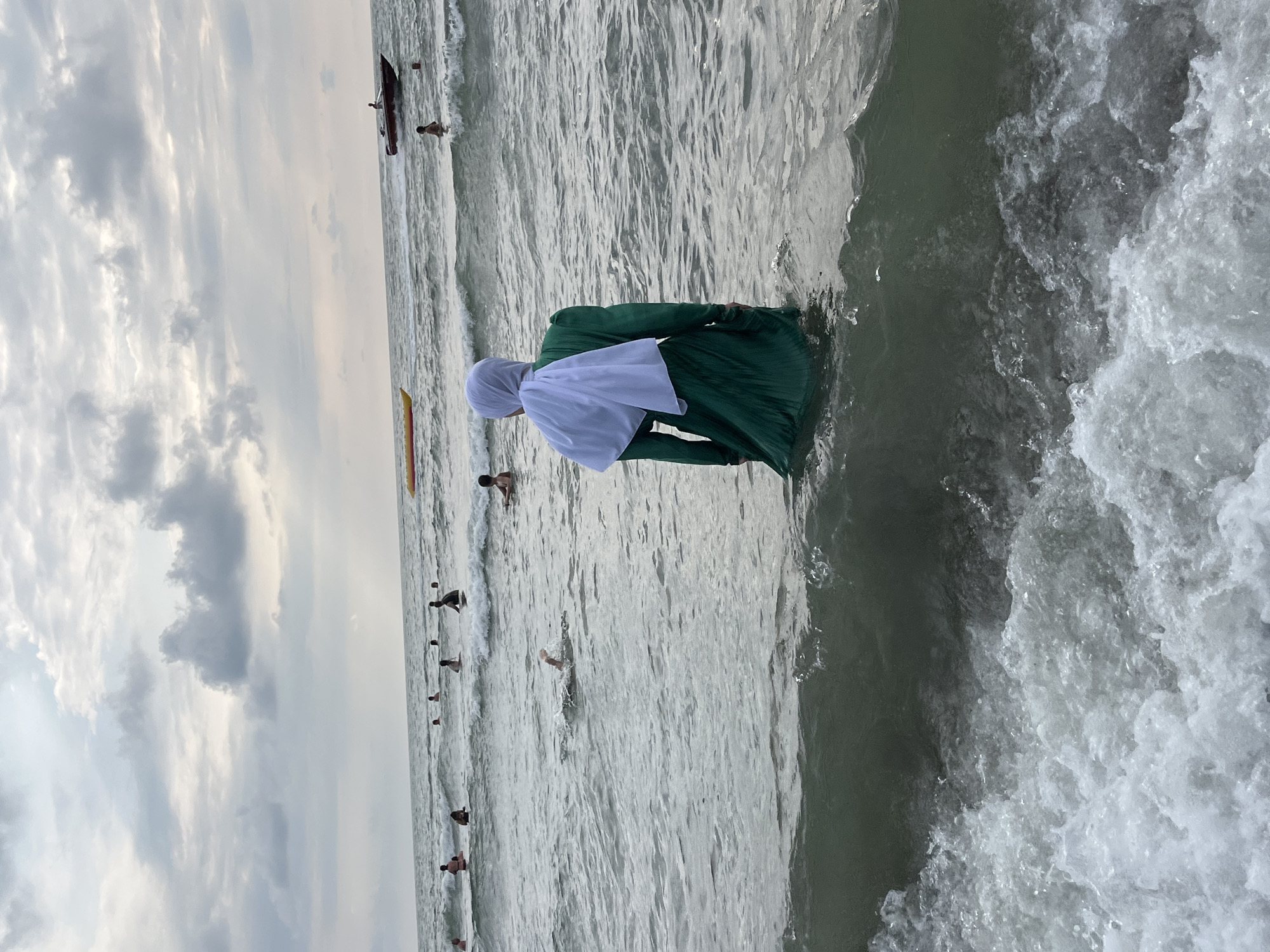 In Ureki, Georgia, a woman wearing a green dress and a white hijab stands in the water at the Black Sea coast. She is facing away from the camera, looking out at the sea where several other people are swimming and wading. The sky is overcast with gray clouds, and the water is moderately choppy with small waves. In the background, a yellow banana boat and a red speedboat are visible on the water, along with other swimmers and bathers spread across the sea. The foreground shows a wave breaking with white foam.