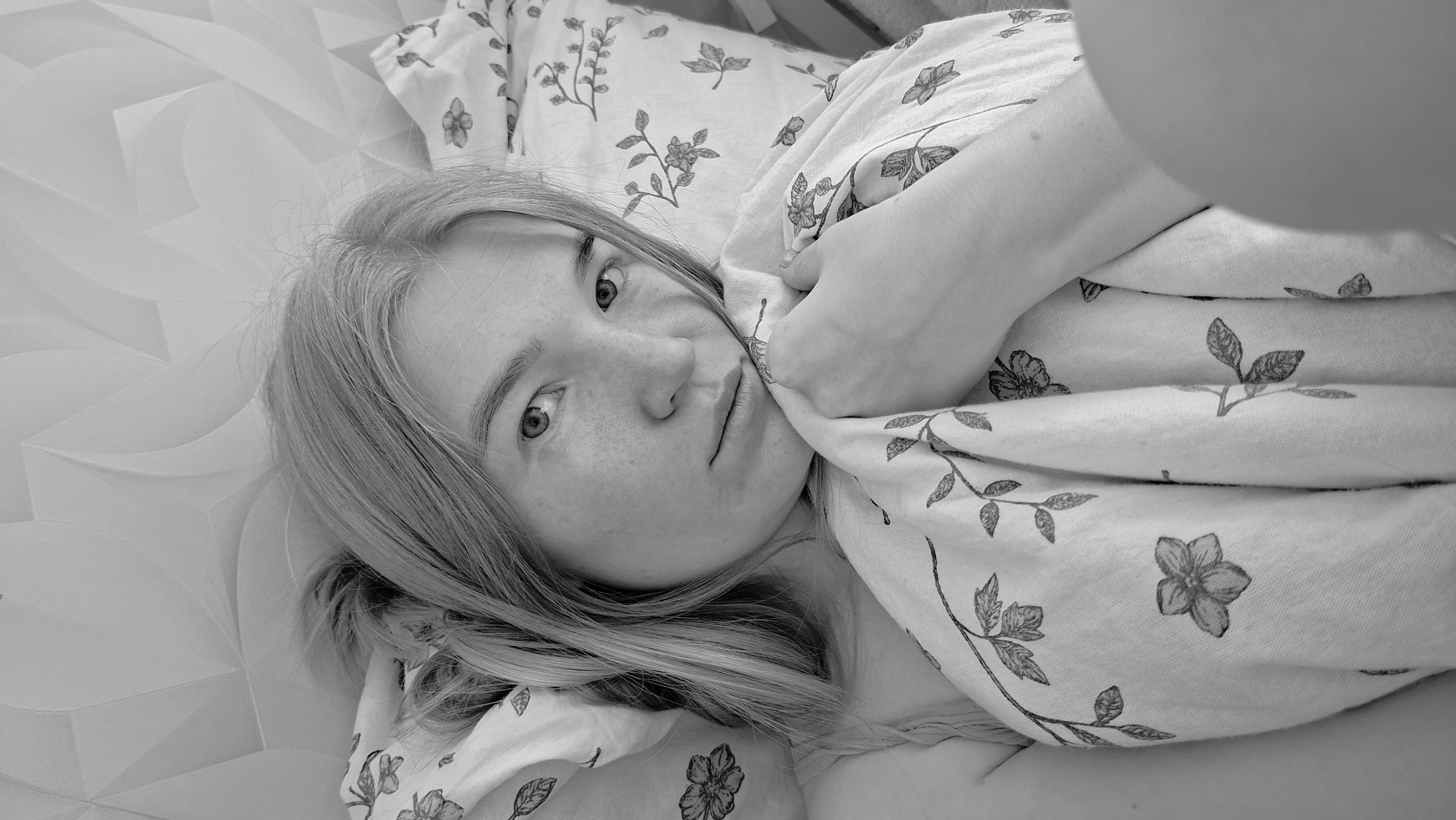 A monochrome, close-up, eye-level shot depicts a young woman lying in bed. She is fair-skinned with freckles, and her long hair is light-colored. She is looking directly at the camera with a slightly concerned expression. She is partially covered by a patterned blanket or sheet featuring botanical illustrations. The bedding beneath her has a geometric, textured pattern.
The location is specified as Chaikovsky, Russia.