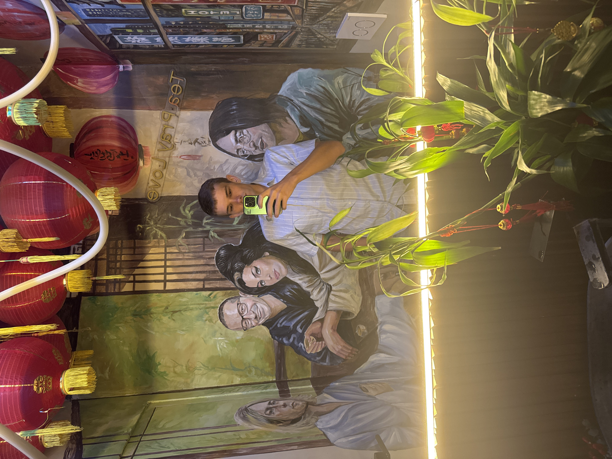 A reflective surface captures a man taking a selfie in what appears to be a restaurant in North Kuta, Indonesia. The reflection includes a wall mural featuring multiple figures in various poses, seemingly in a lush, natural setting, interspersed with red lanterns. A plant stands nearby, partially obscuring the mural and reflecting subject.