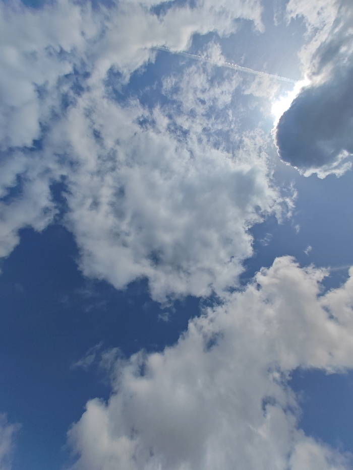 The image depicts a sky scene above Shumen, Bulgaria. The upper two-thirds of the frame show a blue sky partially obscured by a dispersed formation of white and grey cumulus and stratocumulus clouds. The sun is visible in the upper right quadrant, partially occluded by a cloud, producing significant glare and illumination. A distinct, linear white contrail extends diagonally from the upper right towards the center-left. The lower third of the image predominantly features dense white and grey cloud formations. No ground-level features, people, objects, actions, or interactions within the city of Shumen are visible; the scene consists solely of atmospheric elements.