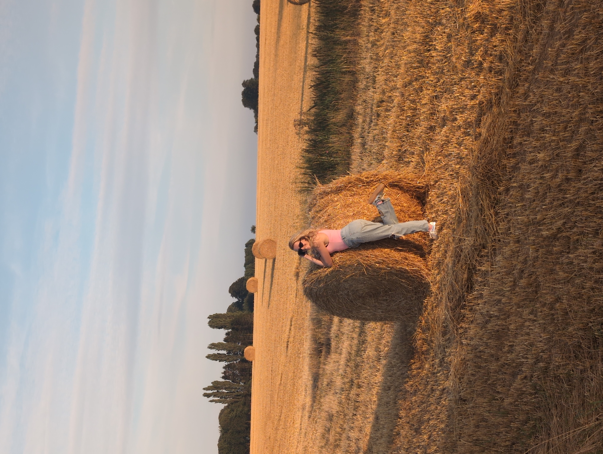 A young woman with blonde hair and sunglasses is posing next to a large, round bale of hay in a wheat field. She is wearing a pink tank top and ripped denim jeans. Her right leg is bent at the knee and extended backward, and she is leaning on the bale with her arms. She appears to be holding a camera to her face, as if taking a photo or being photographed. The field is golden brown, indicating it has been harvested, and several other hay bales are visible in the distance. The sky is a pale blue with wispy clouds, and the sun is casting long shadows across the field, suggesting it is late afternoon or early evening. The scene is set in Alvimare, France.