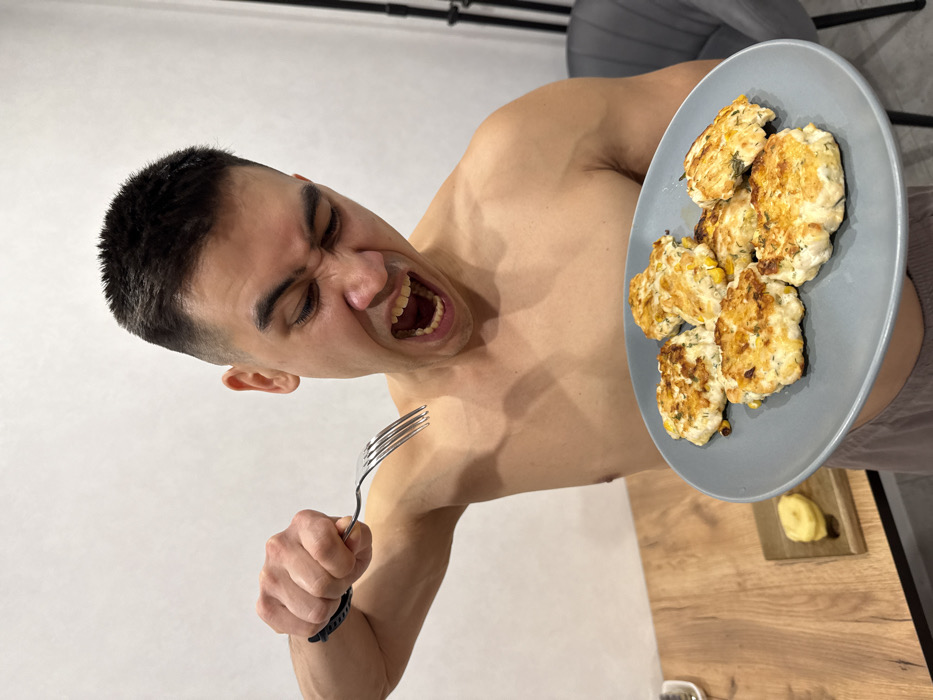 A shirtless man with short dark hair holds a plate of cooked food in one hand and a fork in the other. His eyes are closed, and his mouth is open in what appears to be a grimace or a shout. The plate holds several golden-brown patties, possibly cutlets or fritters. A wooden table and a grey chair are partially visible in the background. A small cutting board with what looks like a piece of fruit sits on the table.