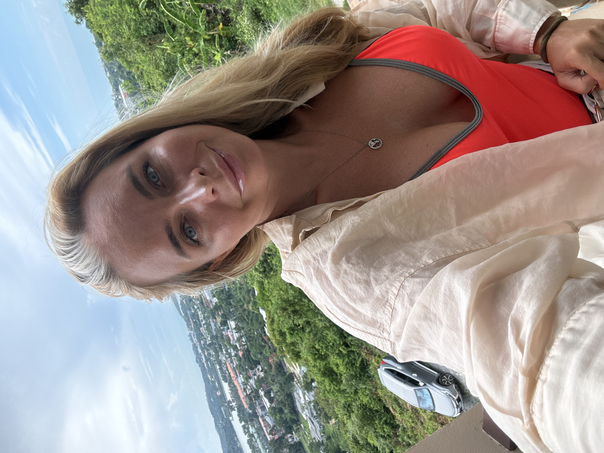 A blonde woman with blue eyes and fair skin is taking a selfie outdoors. She is wearing a bright red swimsuit with grey trim and an open, cream-colored shirt. A silver necklace with a circular pendant hangs around her neck. Her blonde hair is partially pulled back. The background shows a panoramic view of a town or city nestled among lush green hills, with a body of water and the ocean visible in the distance under a partly cloudy sky. A silver car is parked on a nearby road. The woman is smiling and looking towards the camera.