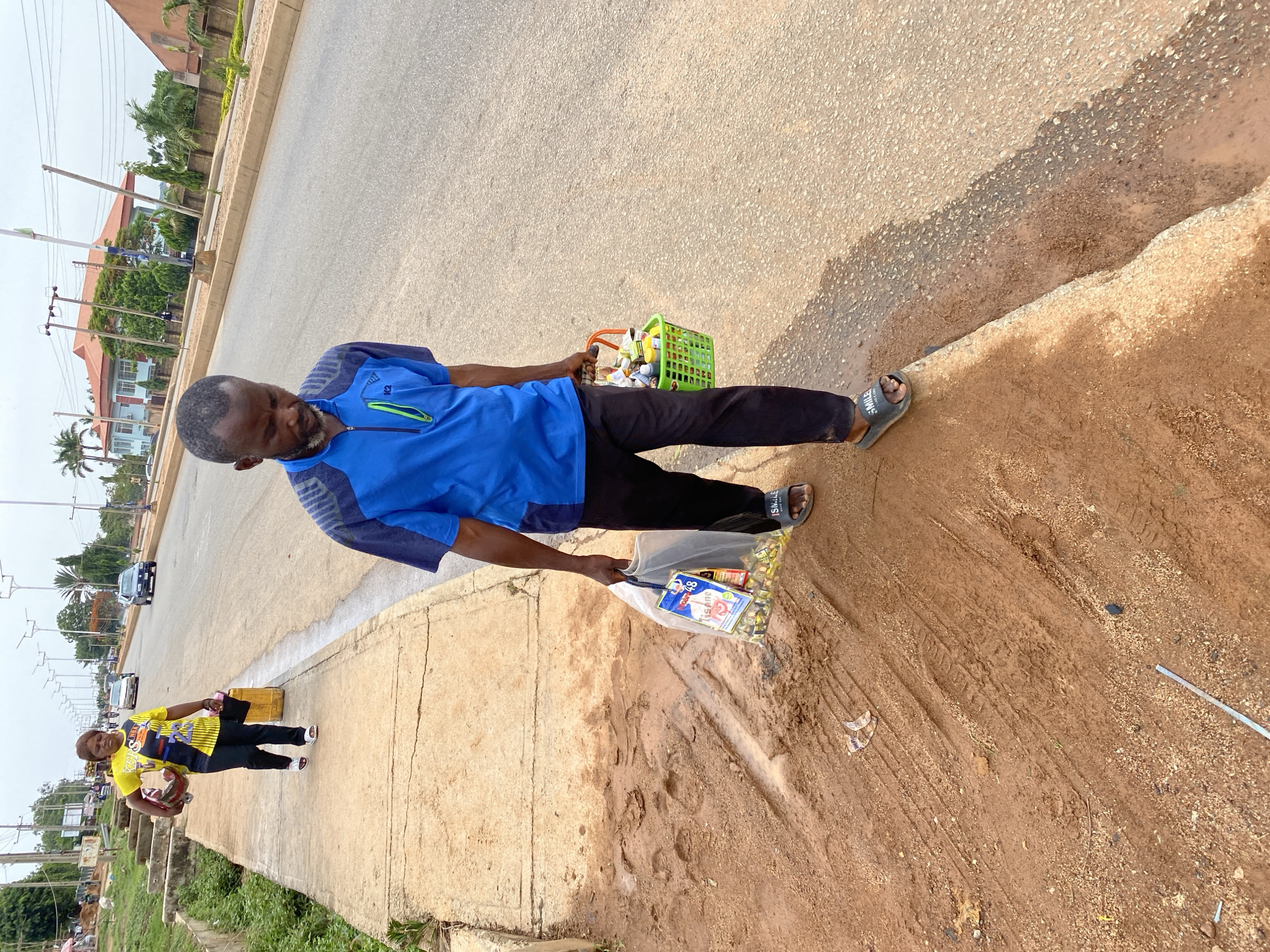 A man stands on a dirt and concrete shoulder of a paved street in Bukuru, Nigeria. He wears a blue collared shirt, black pants, and grey sandals. He carries a green basket in one hand and a clear plastic bag filled with items in the other. In the background, a woman wearing a yellow and black shirt stands on the sidewalk holding a red object. Cars are visible on the paved road. Buildings and trees are also visible in the background.