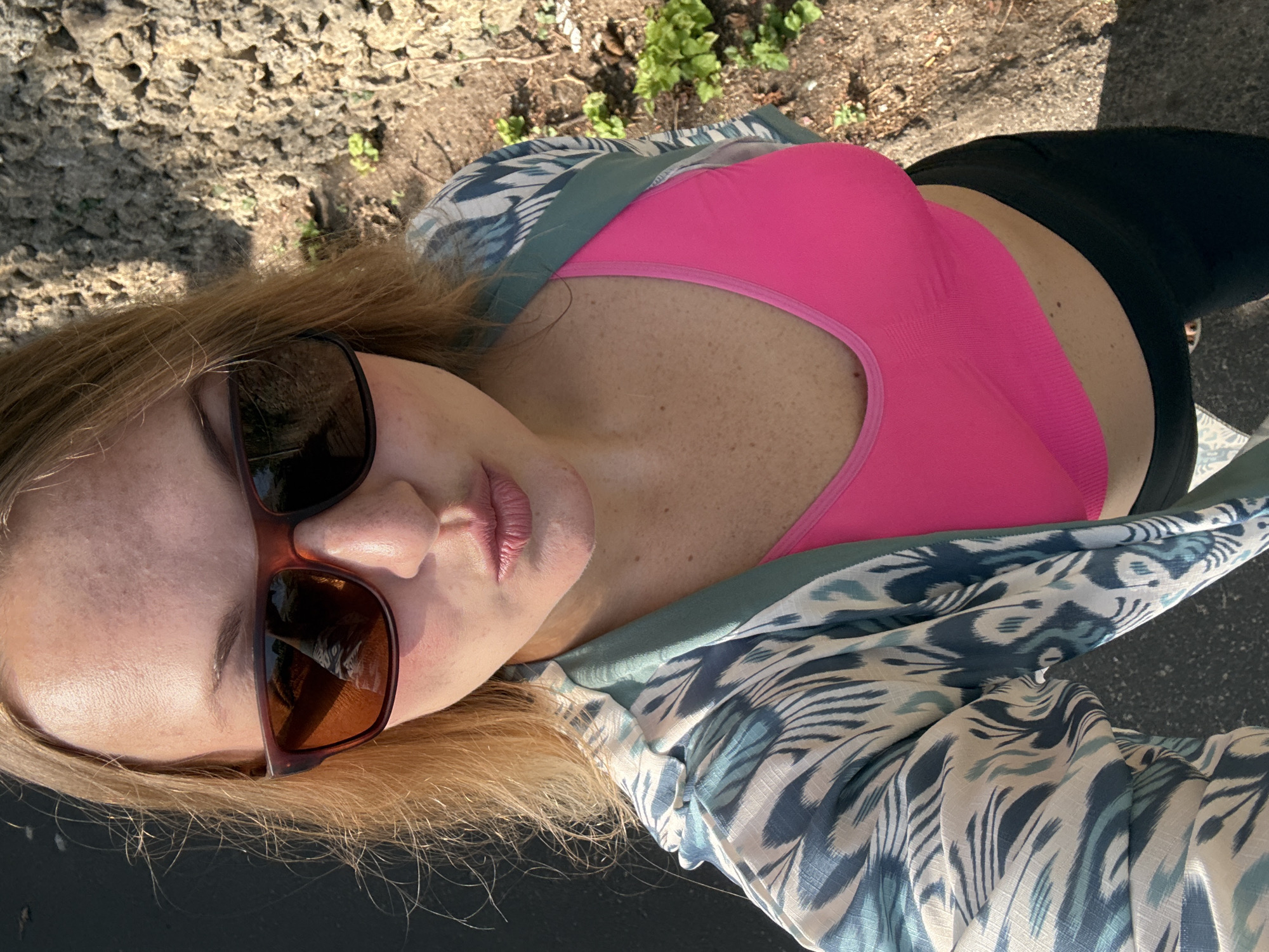 A woman is taking a selfie outdoors, with a natural stone wall and vegetation in the background. She is wearing a bright pink sports bra, a patterned kimono-style jacket with blue and white designs, and black pants. She has on sunglasses with brown lenses. Her lips are pursed in a kiss pose. The scene suggests a warm, sunny day. The overall impression is one of casual outdoor activity.