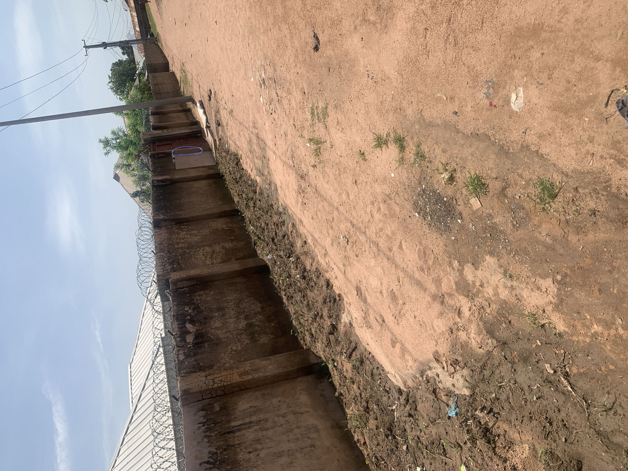 A street scene in Jauro Yinu, Nigeria shows a red dirt road leading into the distance, bordered on the left by a weathered concrete wall topped with barbed wire. The wall appears to enclose a property with a metal roof visible behind it. Sparse green vegetation grows along the base of the wall and sporadically along the edge of the road. The road itself has patches of mud and scattered debris, including plastic bags and paper. A utility pole with electrical wires stands on the right side of the road, further down the street. The sky is bright blue with scattered white clouds. No people are clearly visible in the immediate foreground or midground, though a small figure may be present in the far distance. The overall impression is of an outdoor, semi-urban environment under a clear sky.