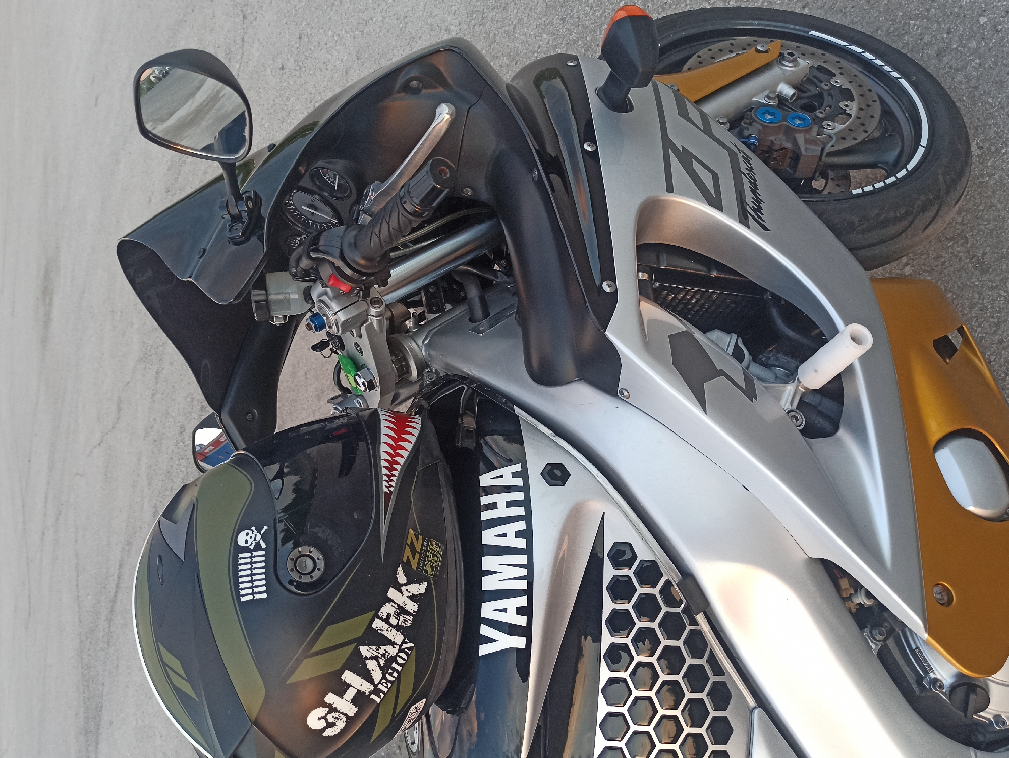 The image depicts a parked Yamaha motorcycle with a helmet resting on the fuel tank. The motorcycle is silver and gold with black accents and "YAMAHA" inscribed on the side. The helmet is black and olive green with a skull and crossbones sticker and "SHARK Legion" inscribed on the side. The front tire of the motorcycle is visible, showing gold-colored forks and blue brake calipers. The location and city are not determinable. There are no people in the image. The environment appears to be a paved surface.