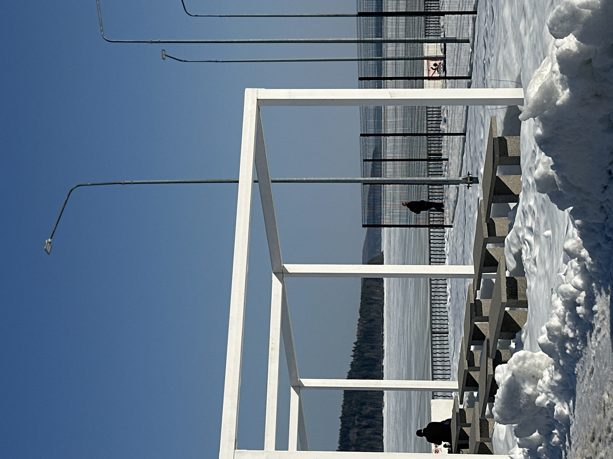The image depicts an outdoor winter scene on a clear, sunny day at a waterfront promenade in Chaikovsky, Russia, likely bordering the Kama Reservoir. In the left and central foreground, a white, open rectangular pergola, constructed from square beams, stands prominently. Beneath and around this structure, approximately seven light grey, rectangular concrete benches or tables are arranged, partially covered by substantial snow accumulation that also forms drifts and piles on the ground, with patches of dark pavement visible.
Two individuals are present: one is seated on a bench under the pergola on the far left, observed from behind, wearing dark winter clothing. A second individual, also in dark clothing, stands further back near a dark metal mesh fence on the right, facing slightly left. This fence, approximately 2 meters in height, extends horizontally across the mid-ground, separating the paved area from a vast, ice-covered body of water. Along this fence line, four slender grey streetlights with upward-curving arms and rectangular LED luminaires are positioned, extending into a cloudless, bright blue sky. On the far side of the frozen water, a dense line of dark coniferous forest is visible along the distant horizon, with faint hills beyond. A white rectangular sign displaying a red "no swimming" symbol is attached to the fence on the right side of the image.