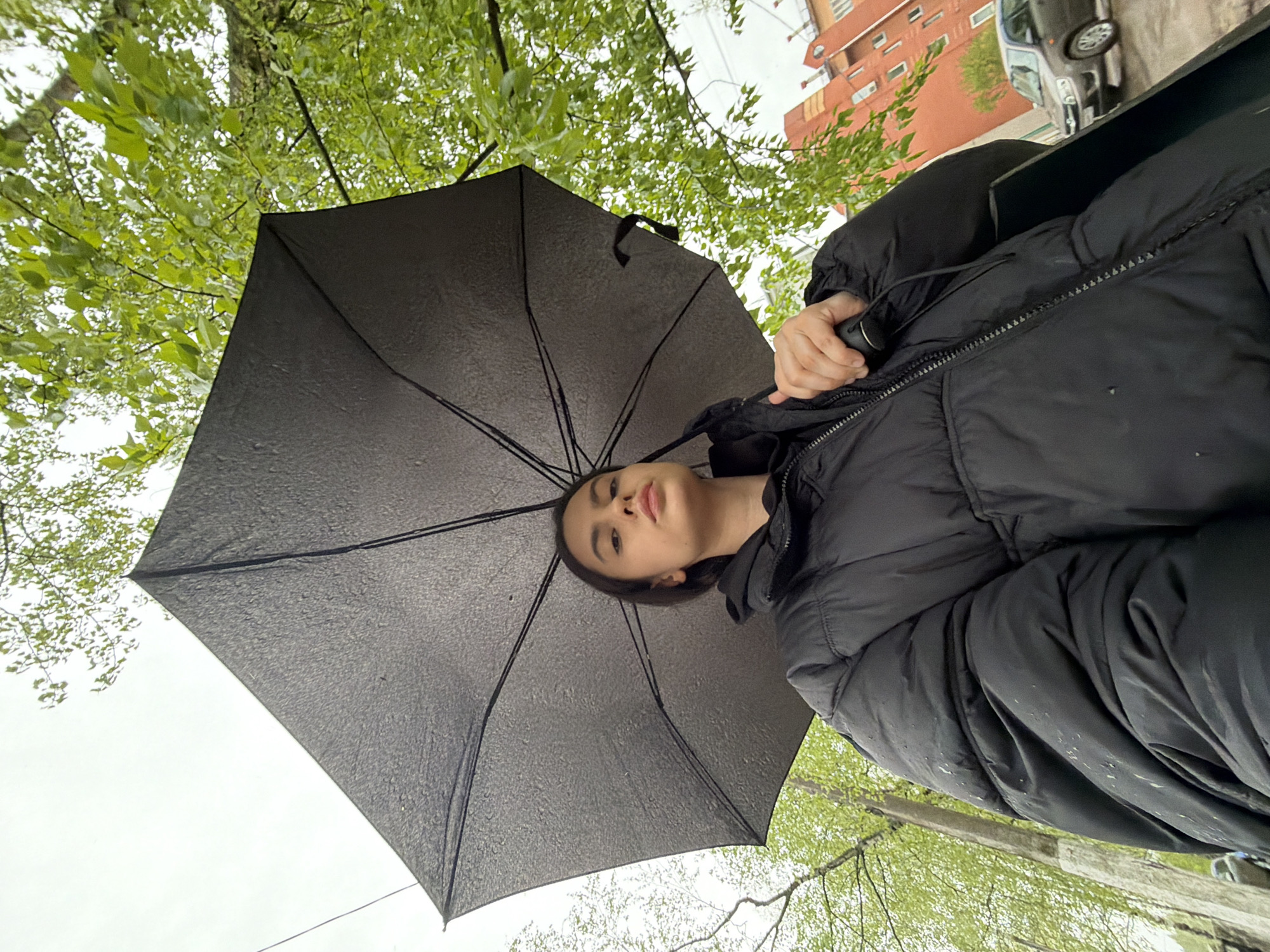 A low-angle shot depicts a woman holding a black umbrella, apparently against rain, under a tree in Chaikovsky, Russia. The umbrella is open and wet, and the woman is wearing a black puffer jacket. In the background are green leaves, buildings, and a parked car.