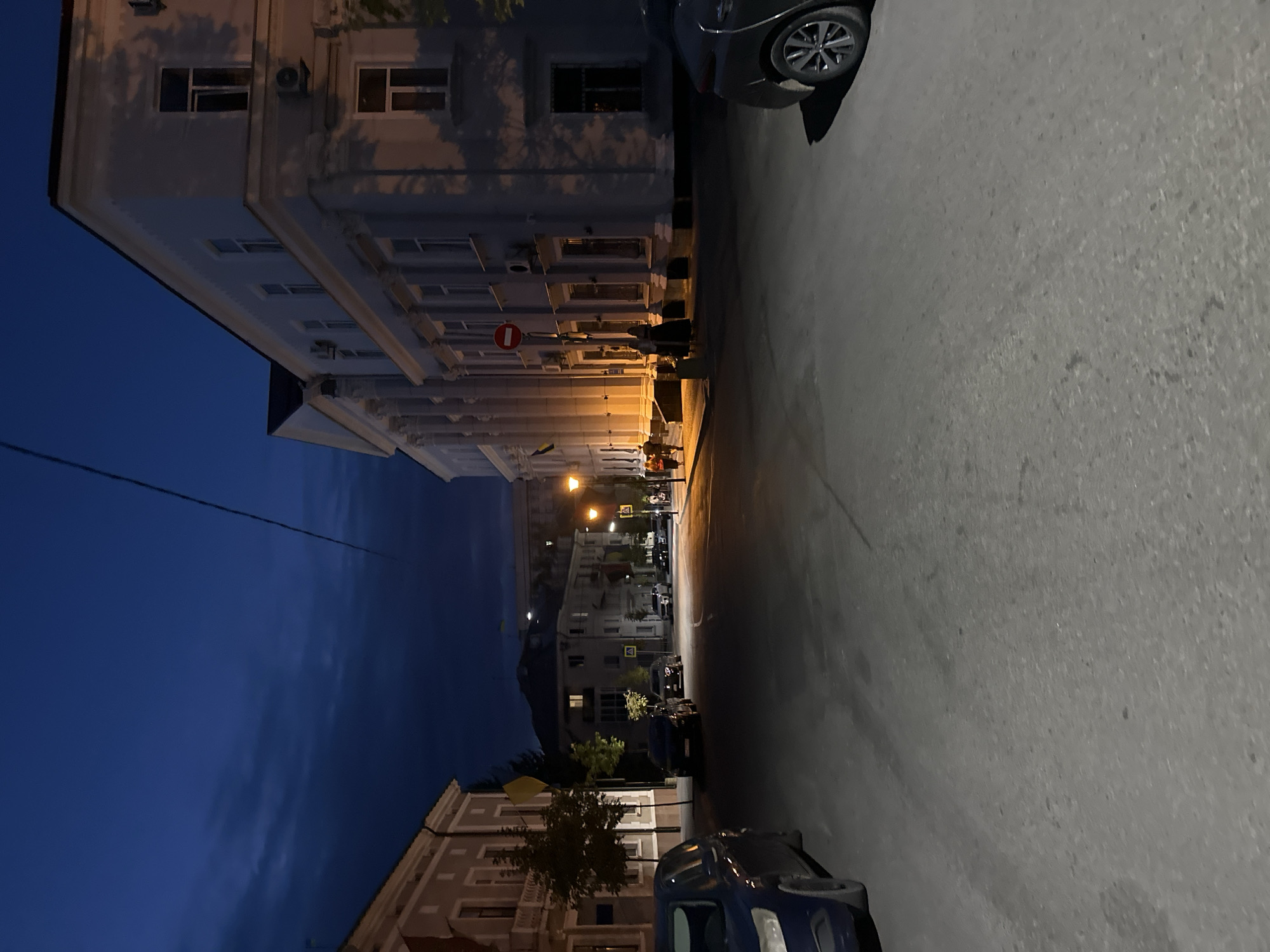 The image depicts a street scene in Ternopil, Ukraine, at dusk or night. The sky is a deep blue with some cloud streaks. On the right side of the street is a large, light-colored building with classical architectural elements, including columns and multiple windows. There are streetlights casting a warm glow on the pavement. A red and white circular sign is visible on the side of the building. Several people are walking on the sidewalk in front of the building, some appearing to be couples. On the left side of the street, there is a lighter-colored building and a streetlamp. Several cars are parked or driving on the street, including a dark-colored SUV in the foreground on the left and a dark-colored sedan on the right. Ukrainian flags are visible on flagpoles in front of some buildings. The overall impression is a quiet, urban street at the end of the day.