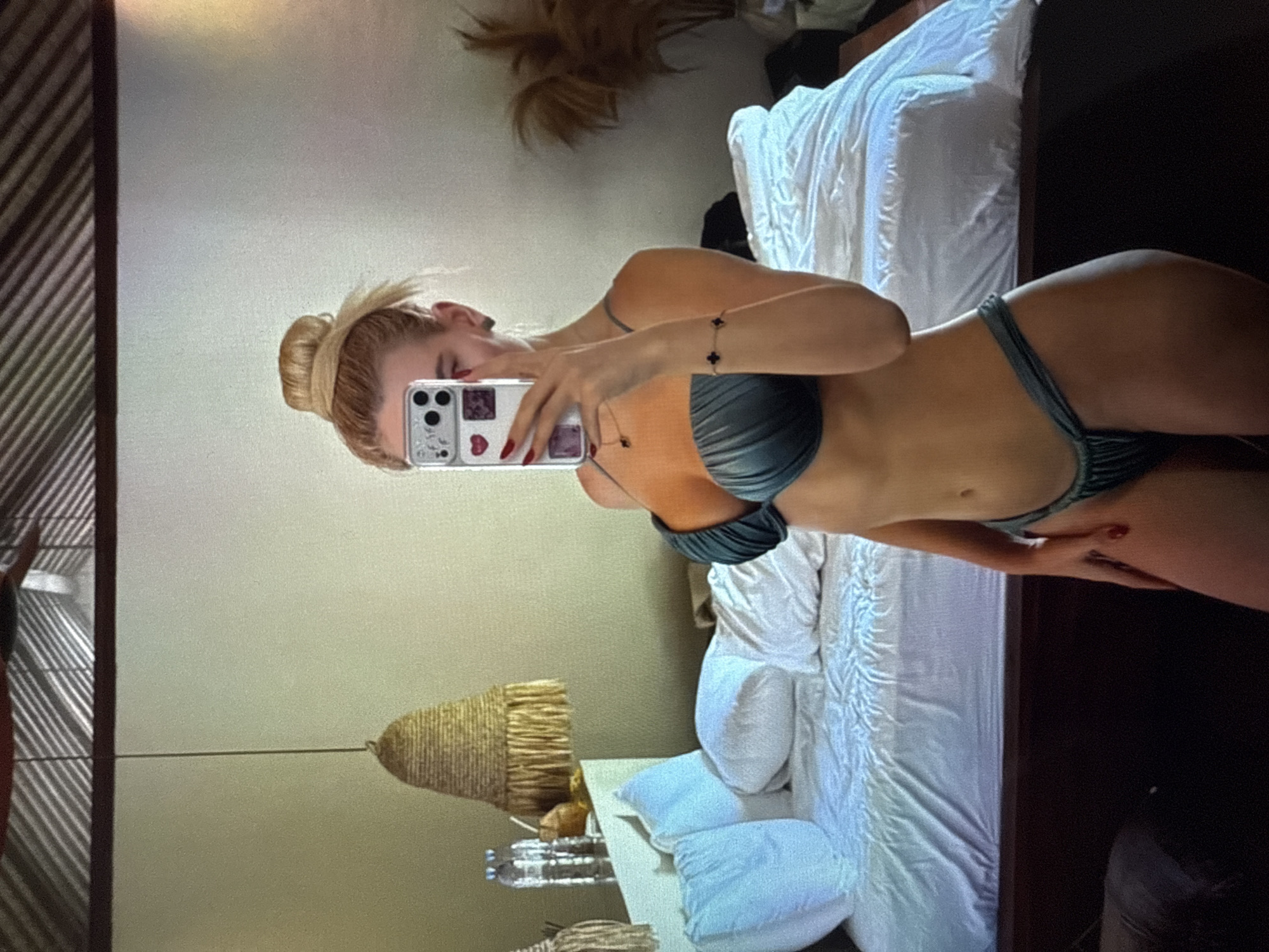 A woman is taking a mirror selfie in a bedroom. She is wearing a teal bikini and her hair is pulled up in a bun. The bed in the background is unmade, with white sheets and pillows. There are two bottles of water and a coconut drink on a nightstand next to the bed. A woven lampshade hangs from the ceiling. The room has a neutral color palette with light beige walls.