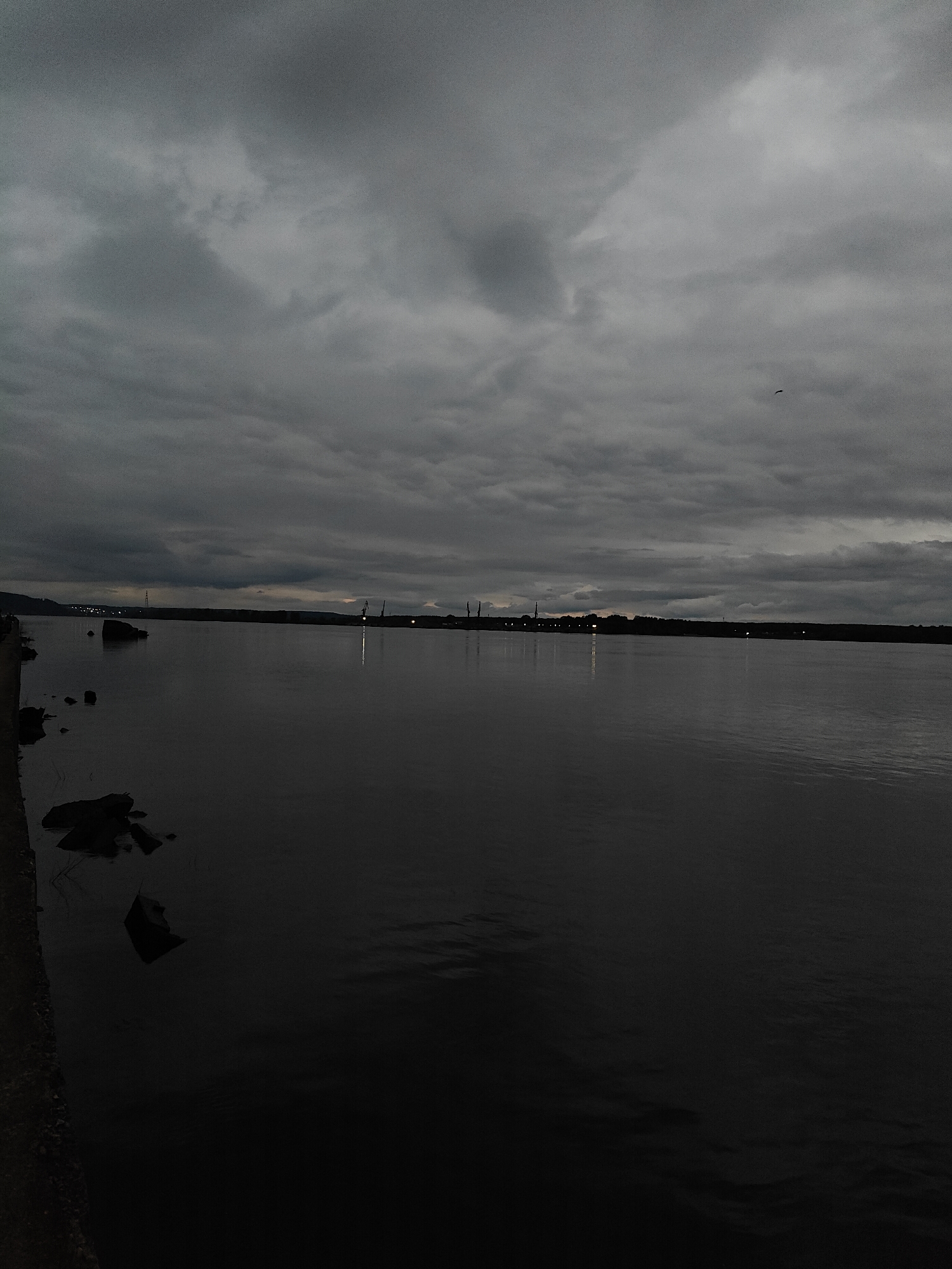 The image shows a body of water, likely a river or lake, under a heavily clouded, overcast sky. The water is dark and calm, reflecting the dim light from the sky. On the left edge of the image, there is a shoreline with rocks and what appears to be a concrete edge. Across the water, there is a distant horizon with some indistinct structures that appear industrial, including possible cranes or towers. There is also a single bird in the sky. The location could be Chaikovsky, Russia. There are no people visible.