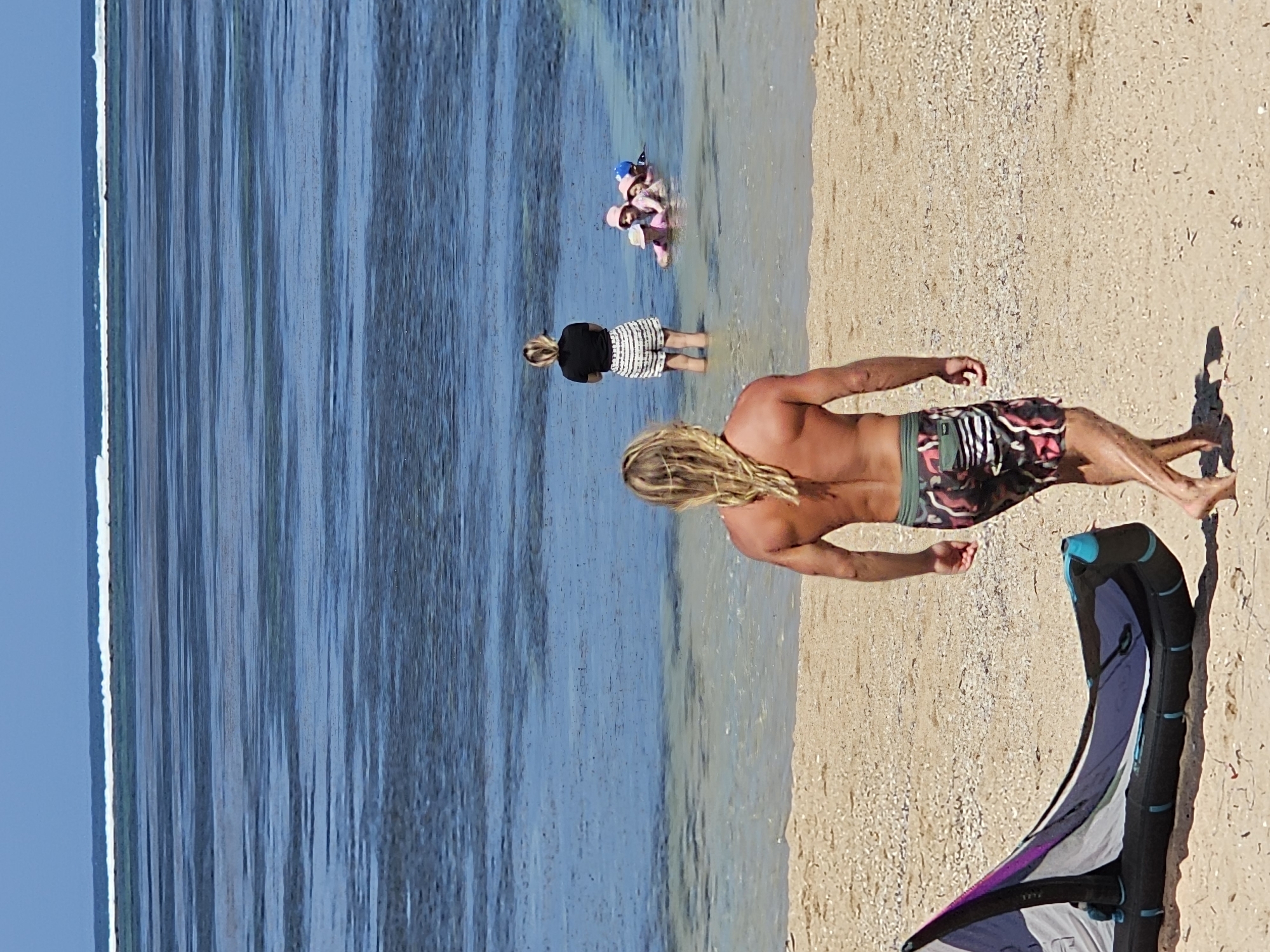 The image depicts a beach scene with people in the water and on the shore. In the foreground, a man with long blonde hair and a muscular build is walking away from the camera towards the water. He is wearing patterned swim trunks. To his left on the sand is a deflated kite, partially visible. In the shallow water, a woman is standing, facing the group of children in the water. Three young children, wearing pink hats and swim caps, are playing in the water. The water is shallow and appears to be calm with visible ripples. The background shows a calm ocean with a distinct line of breaking waves in the distance. The beach is composed of light-colored sand and small shells. The overall impression is a bright, sunny day at a tropical beach. The scene is consistent with a location in Denpasar, Indonesia, known for its beaches and water sports.