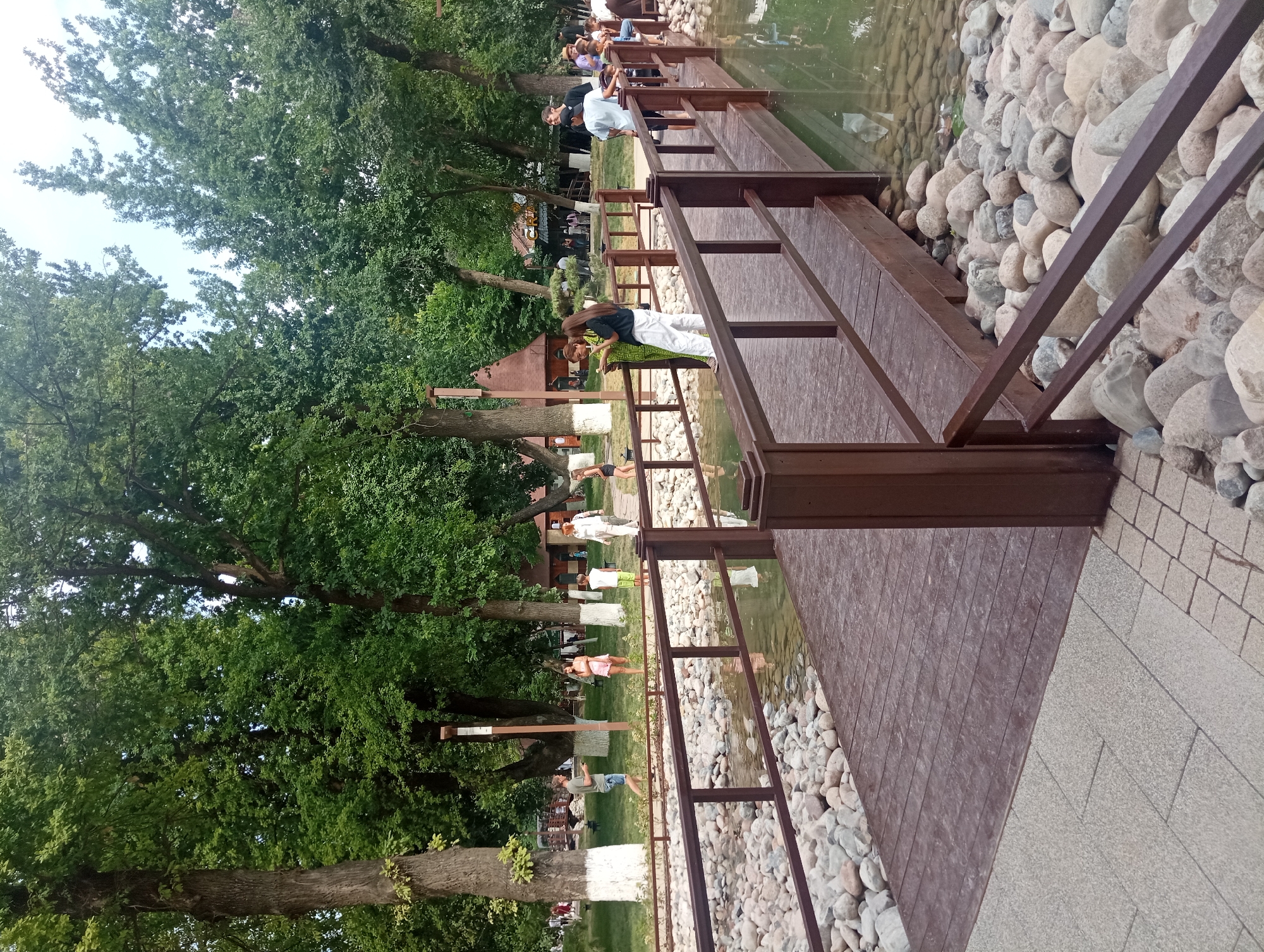 A wooden footbridge crosses a shallow body of water filled with smooth, rounded stones. Several individuals are scattered across the bridge and the surrounding grassy area. To the left, a man wearing a green t-shirt and blue shorts walks away from the bridge, looking at his phone. A woman in a pink dress stands near him, looking towards the bridge. Further back, a child in a green shirt and shorts stands near a pathway. On the bridge, a woman in a green and yellow patterned outfit is engaged in conversation with a woman in a black shirt and white pants. Another group of people are visible further down the bridge, some standing and talking. In the background, a wooden structure with a conical roof is nestled amongst large, leafy trees. The scene appears to be a park or recreational area within the city of Shymkent, Kazakhstan, characterized by lush greenery and a tranquil water feature.
