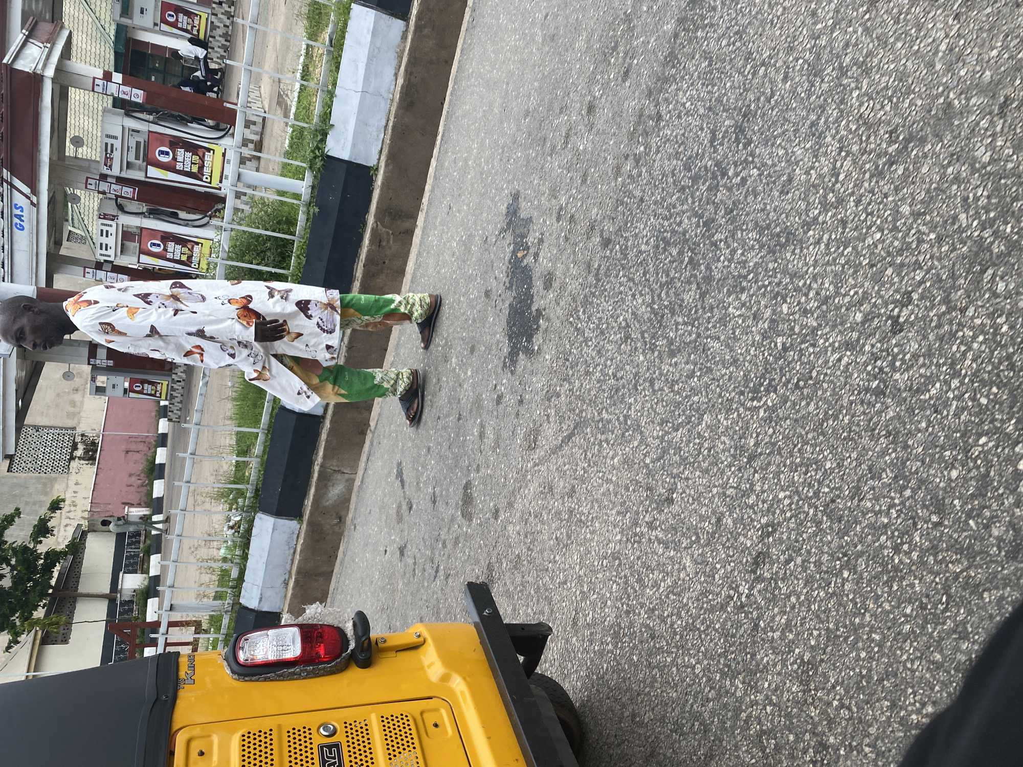A man walks past a gas station in Jalingo, Nigeria. He is wearing a white robe with a butterfly pattern and green pants. A yellow auto-rickshaw is parked to his left. The gas station has multiple pumps and a "GAS" sign. The ground is paved with asphalt.