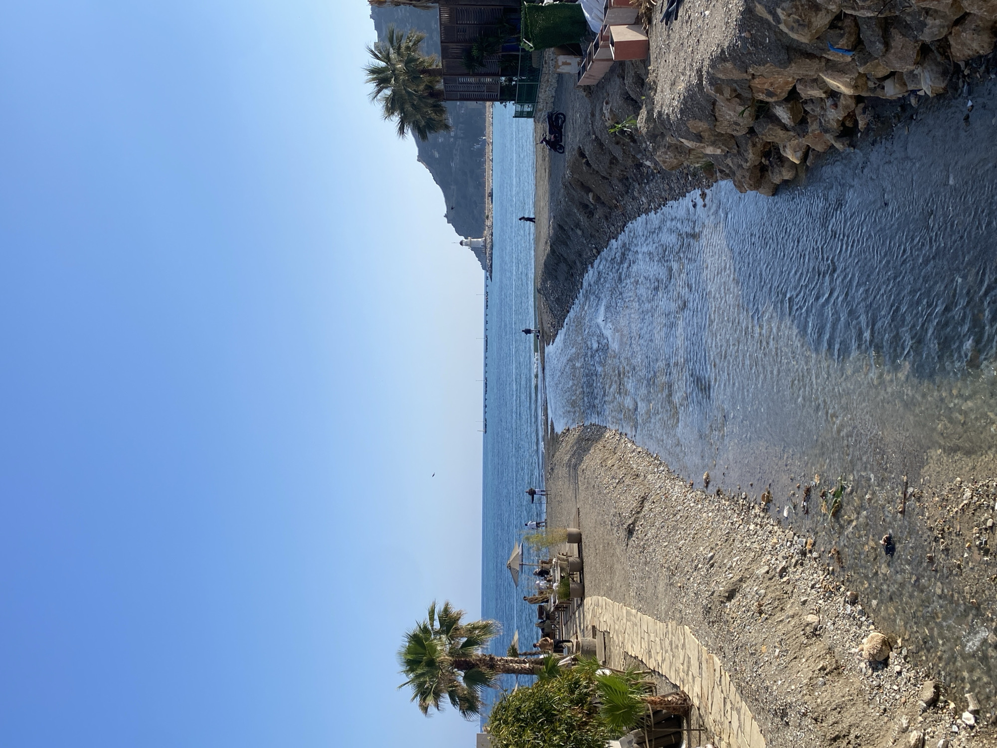 This image, captured in a vertical orientation but depicting a horizontal landscape, showcases a sun-drenched coastal scene in Alanya, Turkey. The upper portion of the frame is dominated by a vast, clear blue sky, indicating bright and pleasant weather, likely during the mid-morning or afternoon.
Below the sky, the calm, deep blue waters of the Mediterranean Sea stretch towards the horizon. In the distance, a distinctive rocky headland juts into the sea, topped by a white lighthouse or a similar prominent structure. Several small, faint boats are discernible far out on the water.
In the foreground and middle ground, a narrow stream of clear water flows from the right side of the image, meandering across a pebbly and sandy beach before emptying into the sea. The stream's immediate right bank is formed by rugged, stacked rocks and earth, with some small green plants. Further back on this bank, behind a low retaining wall, two motorcycles are parked near some rustic wooden and concrete structures.
On the stream's left bank, a wider expanse of pebbly beach gives way to a paved walkway or patio area in the lower left foreground. This area is furnished with several outdoor tables and chairs, some shaded by light-colored umbrellas, suggesting a beachside cafe or restaurant. A few individuals are seated at these tables, relaxing. The area is adorned with lush green vegetation, including several prominent palm trees with their characteristic fronds.
Throughout the scene, several people are present. Individuals are scattered along the shoreline, some standing near the water's edge where the stream meets the sea, possibly engaged in leisure activities or fishing. The overall atmosphere is one of tranquility and enjoyment of the sunny coastal environment. No visible text is present in the image.