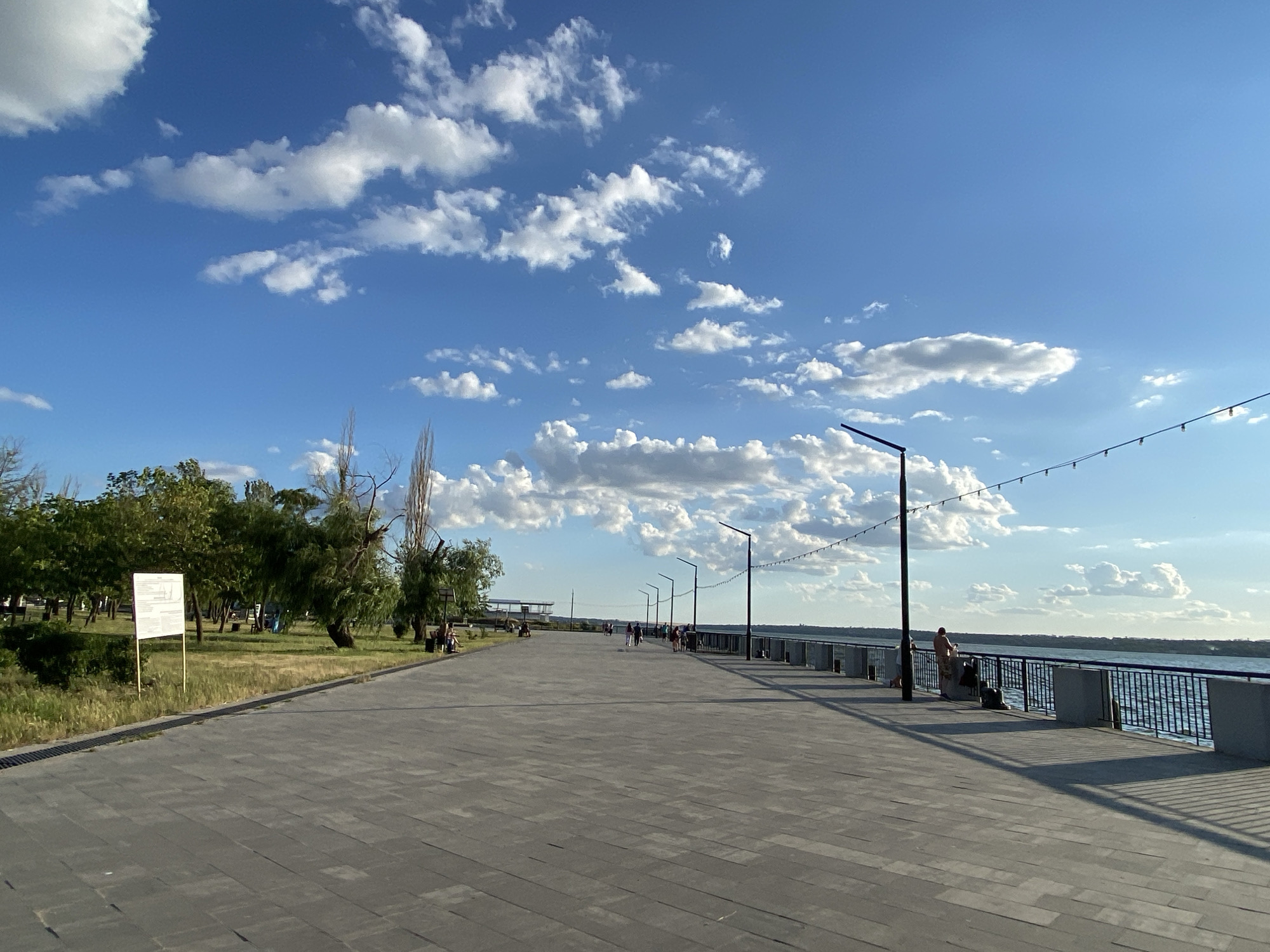A wide, paved promenade stretches into the distance along a body of water under a blue sky dotted with white clouds. The promenade is lined with streetlights and a string of decorative lights. On the left side of the promenade, there is a grassy area with trees and benches. Several people are visible walking along the promenade, and a few are seated on benches. On the right side, a railing separates the promenade from the water. The scene appears to be a public park or recreational area.