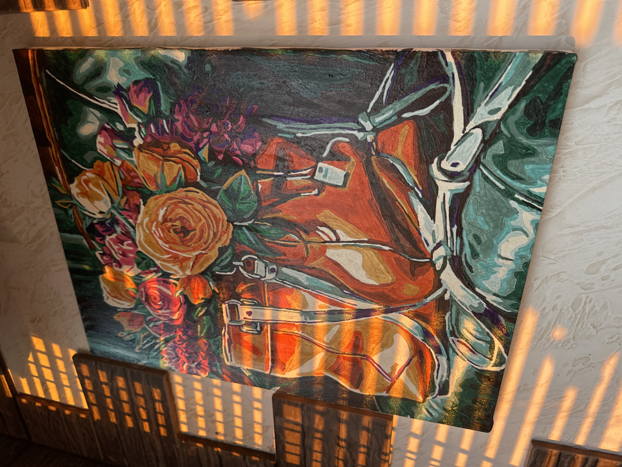 The media file presents a vibrant painting, likely an acrylic or oil on canvas, depicting a colorful floral arrangement next to what appears to be a stylized brown and orange leather bag or saddlebag. The bouquet features prominent orange, yellow, and red roses, interspersed with purple and pink blossoms and green leaves, all set against a dark background with teal and turquoise accents. The painting exhibits a distinct style with bold outlines and color blocking, reminiscent of a paint-by-numbers approach.
The painting is situated indoors, leaning against or mounted on a light-colored, textured wall. Strong, warm, golden sunlight streams into the room, casting distinct, parallel striped shadows across the wall, the painting's surface, and a wooden structure (possibly a chair back or railing) visible at the bottom. This dramatic lighting suggests either early morning or late afternoon, commonly known as the golden hour. No specific activity is depicted in this still life scene. The image contains no visible text. The location is Tomilino, Russia.