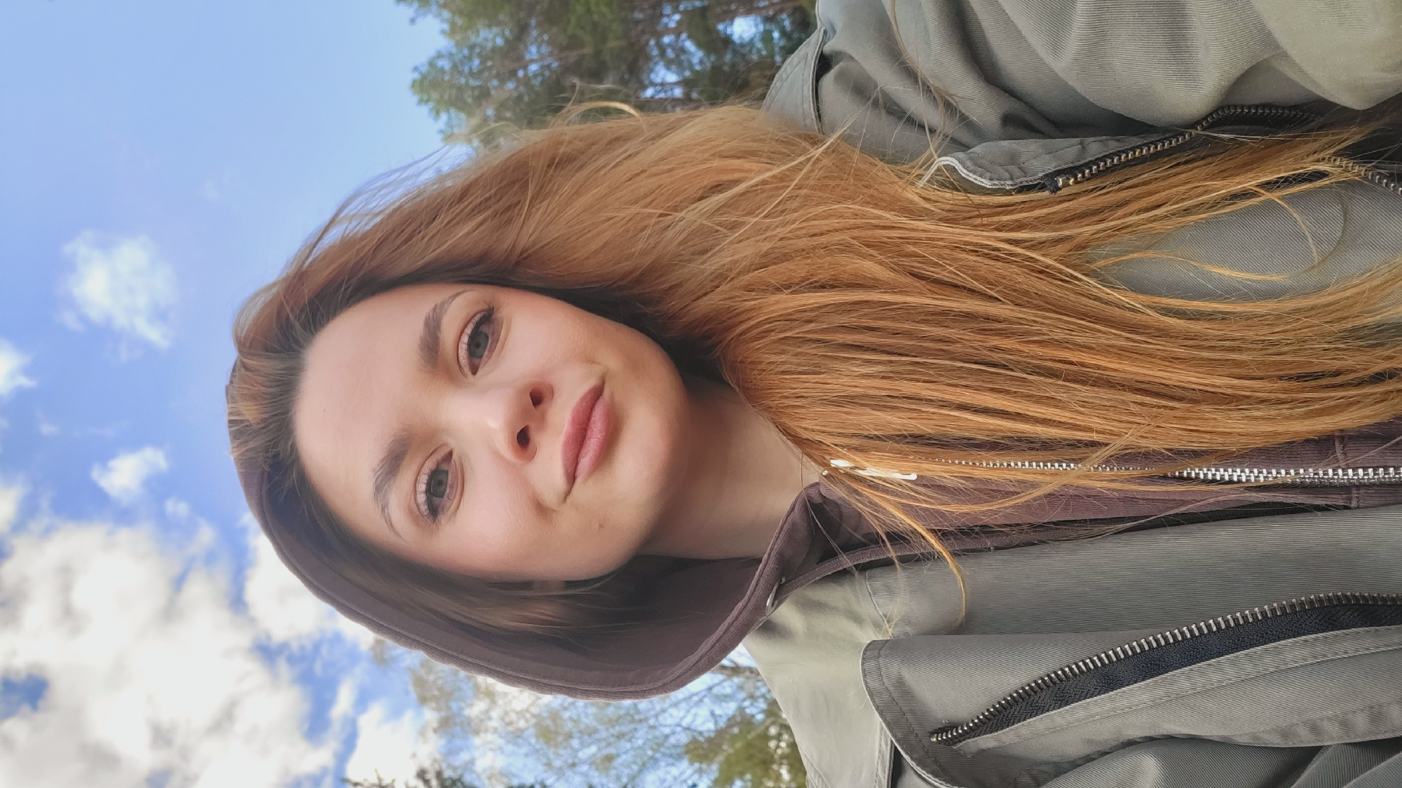 A fair-skinned woman with long, reddish-blonde hair is looking directly into the camera, tilted at an angle, appearing to be taking a selfie. She has light eyes, a small mole on her cheek, and is wearing minimal makeup. She is wearing a hooded jacket with a brown inner layer and a green outer layer. The zipper of the jacket is visible. Behind her is a blue sky with scattered white clouds and the blurred background of trees. Without more data, the location in Chaikovsky, Russia cannot be verified.