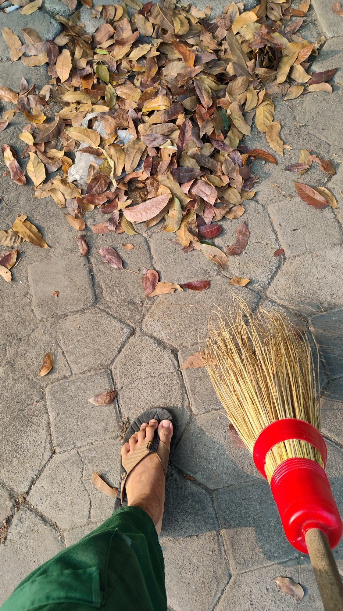 A first-person perspective shot shows a person's left foot wearing a brown sandal and dark green pants, standing on a paved walkway in Pabelan, Indonesia. The ground is covered with a pile of dried brown leaves. A broom with a bright red handle and natural straw bristles is positioned to the right of the foot, suggesting an action of sweeping or the immediate aftermath of sweeping. The paved surface is composed of interlocking hexagonal and rectangular stone tiles. The lighting suggests it is daytime, with shadows indicating the sun is at an angle.