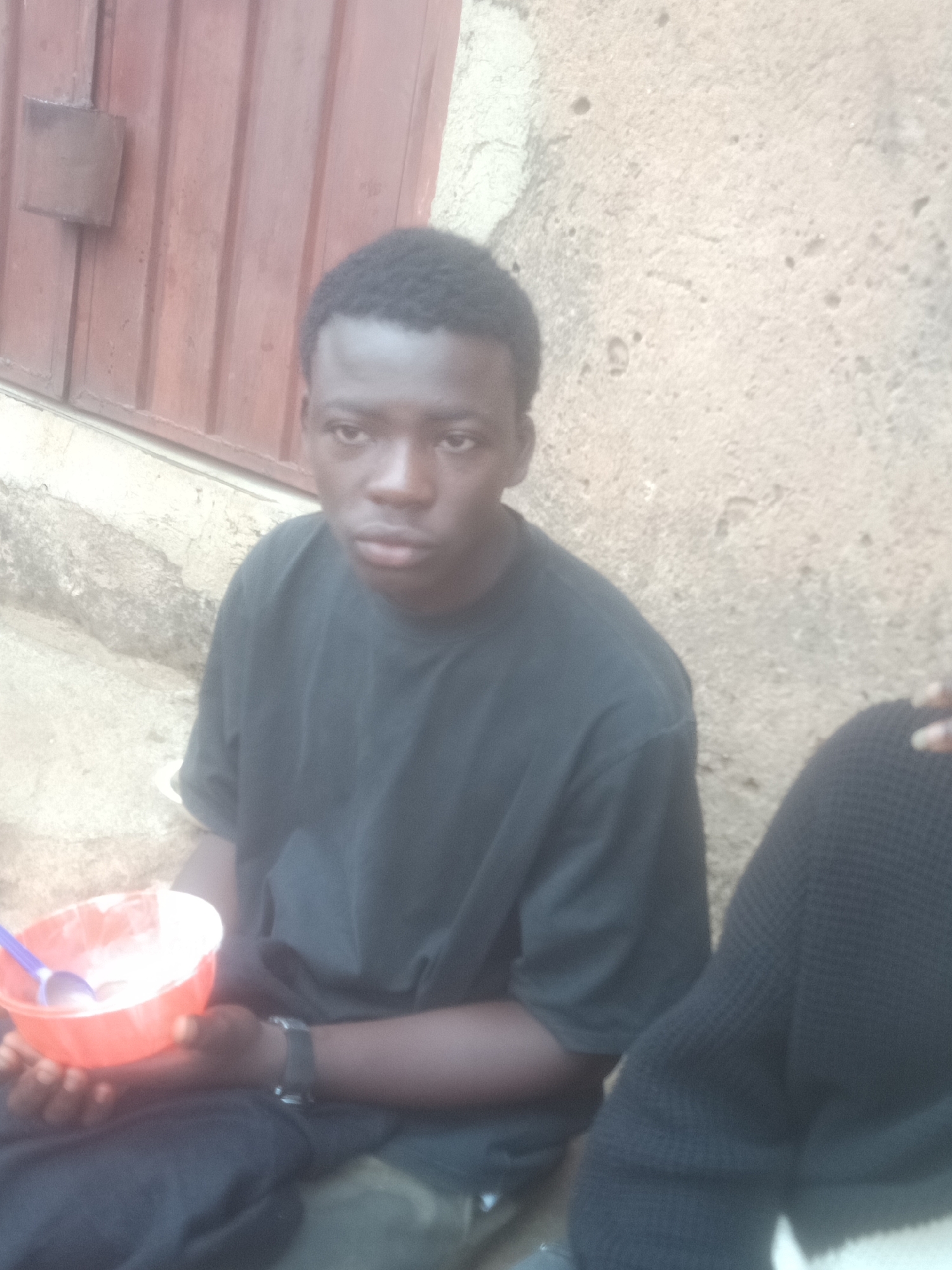 A young Black male is seated outdoors, holding a red bowl containing a white substance, possibly food, and a purple spoon. He is wearing a black t-shirt and black pants, and a watch on his left wrist. The background features a textured concrete wall and a red wooden door or panel. The scene appears to be in an urban environment, though the specific city and location are unknown.