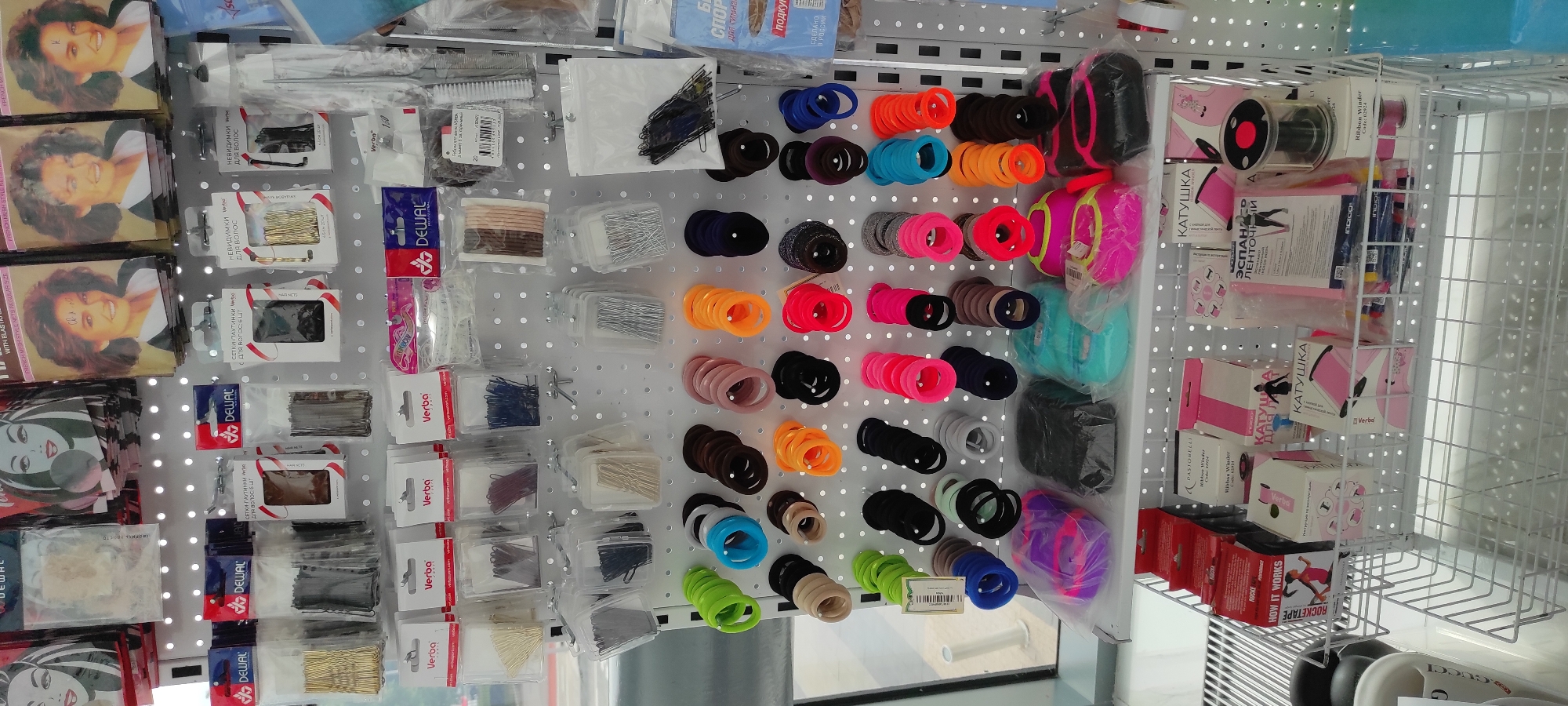 The image shows a display shelf in a store, likely a beauty supply or general merchandise store, filled with various hair accessories and other small items. The shelf is white and perforated, with items hung on hooks or placed in small packages.
On the upper shelves, there are packages of hairnets, hair clips, bobby pins, and hair ties in various colors and sizes. Some packages have branding such as "DEWAL" and "Verba." There are also some cards with images of women with different hairstyles.
The middle shelves are primarily filled with hair ties. These are arranged in rows and columns, with a wide range of colors including green, blue, black, brown, pink, orange, and red. Some are plain, while others appear to have a textured or glittery finish.
The bottom shelf contains larger packaged items. On the left, there are red and black boxes, one with the text "ROCKETAPE" and an illustration of a woman running. Further to the right, there are pink and white boxes, some with the text "КАТУШКА" (which means "spool" or "reel" in Russian). There are also some transparent plastic packages with coiled items inside, and a clear plastic container with a red and black lid, possibly for storing ribbon or thread. Below these shelves, a white wire basket is visible.
The overall environment suggests an indoor retail setting. The lighting appears to be artificial, typical of store lighting. There are no people visible in the image. The items are neatly arranged, indicating a well-stocked and organized display. The presence of Russian text on some of the packaging suggests the location is likely in Russia, and based on the prompt's constraint, it is in Moscow.
