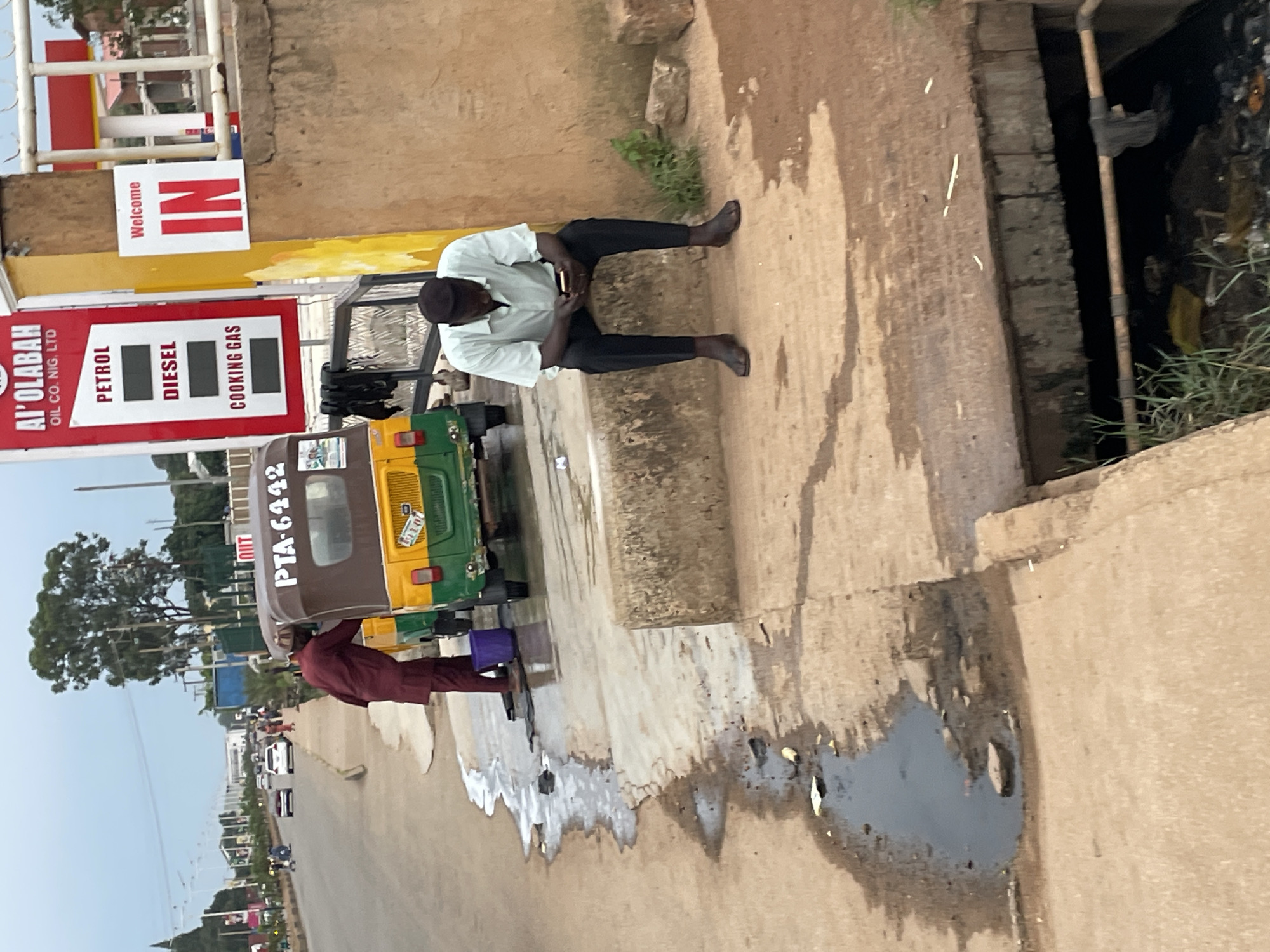 In Barakin Lerawi, Nigeria, a man in a maroon traditional outfit is washing a yellow and green auto-rickshaw by the side of a street. He is standing near a purple bucket and a dark mat on the ground, with soapy water and puddles on the concrete. Further down, a man in a light blue short-sleeved shirt and dark pants is sitting on a concrete ledge, looking at his phone. Behind the auto-rickshaw, a red and white sign for "AI' OLABAH OIL CO. NIG. LTD" is visible, advertising petrol, diesel, and cooking gas, along with a yellow sign that says "Welcome IN". The environment appears to be a roadside with some greenery and a drainage ditch filled with debris. The sky is overcast.