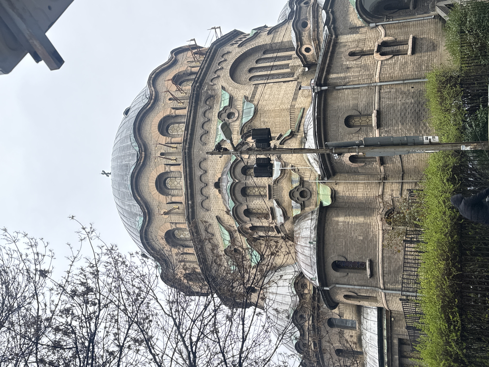 The image depicts the exterior of the St. Alexander Nevsky Cathedral in Sofia, Bulgaria, viewed from below and slightly to the side. The focus is on the dome and upper levels of the building, showcasing its architectural features, including layered dome sections, arched windows, and decorative elements. A section of a power pole is visible to the right side of the shot. Some bare trees are visible to the left of the structure. The sky is overcast, giving the building a muted tone. A line of greenery appears to the right of the structure as well.