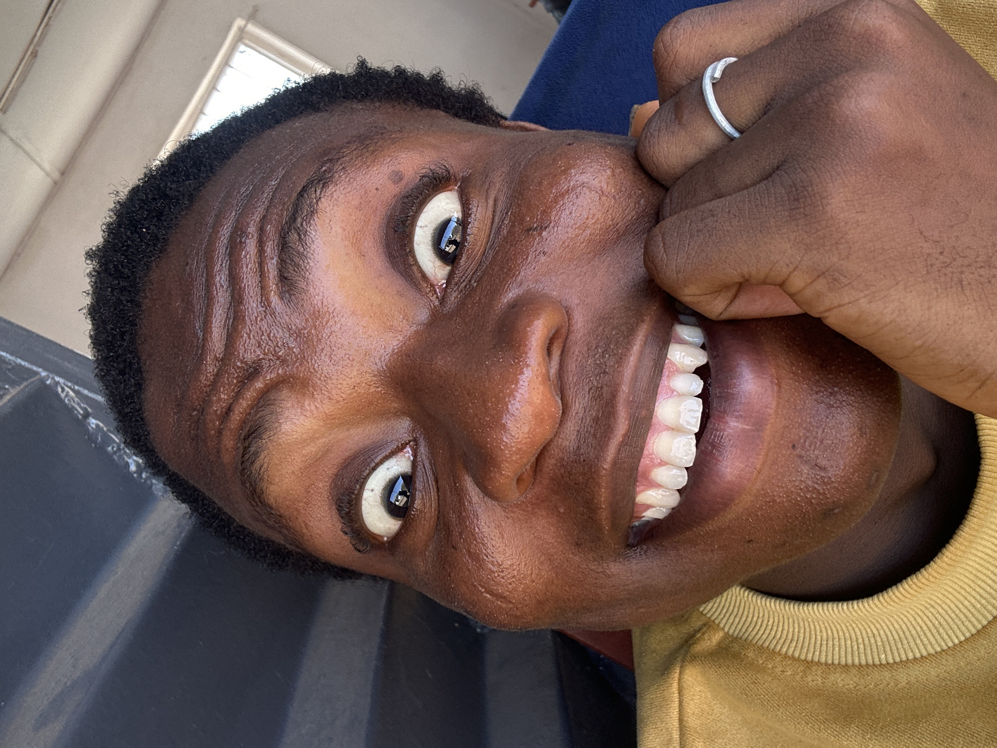 A close-up, rotated view captures a person with dark skin and short, dark hair, smiling broadly to reveal white teeth. The person's left hand, adorned with a silver ring, is partially visible near their cheek. The person is wearing a yellow top and appears to be inside a building. The location is Unknown, Unknown.