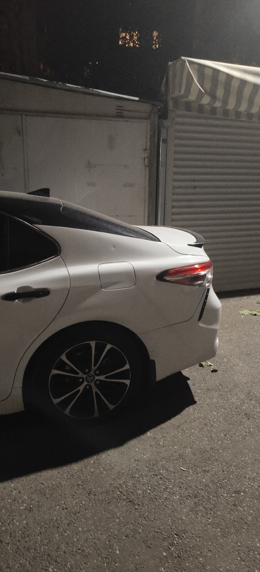 A white sedan is parked on an asphalt surface in front of a garage and a striped awning. The car is viewed from the rear passenger side, showing the tail light, wheel, and part of the trunk. Dust covers the roof and trunk of the car. In the background, a building with illuminated windows is visible. The scene appears to be in an urban environment.