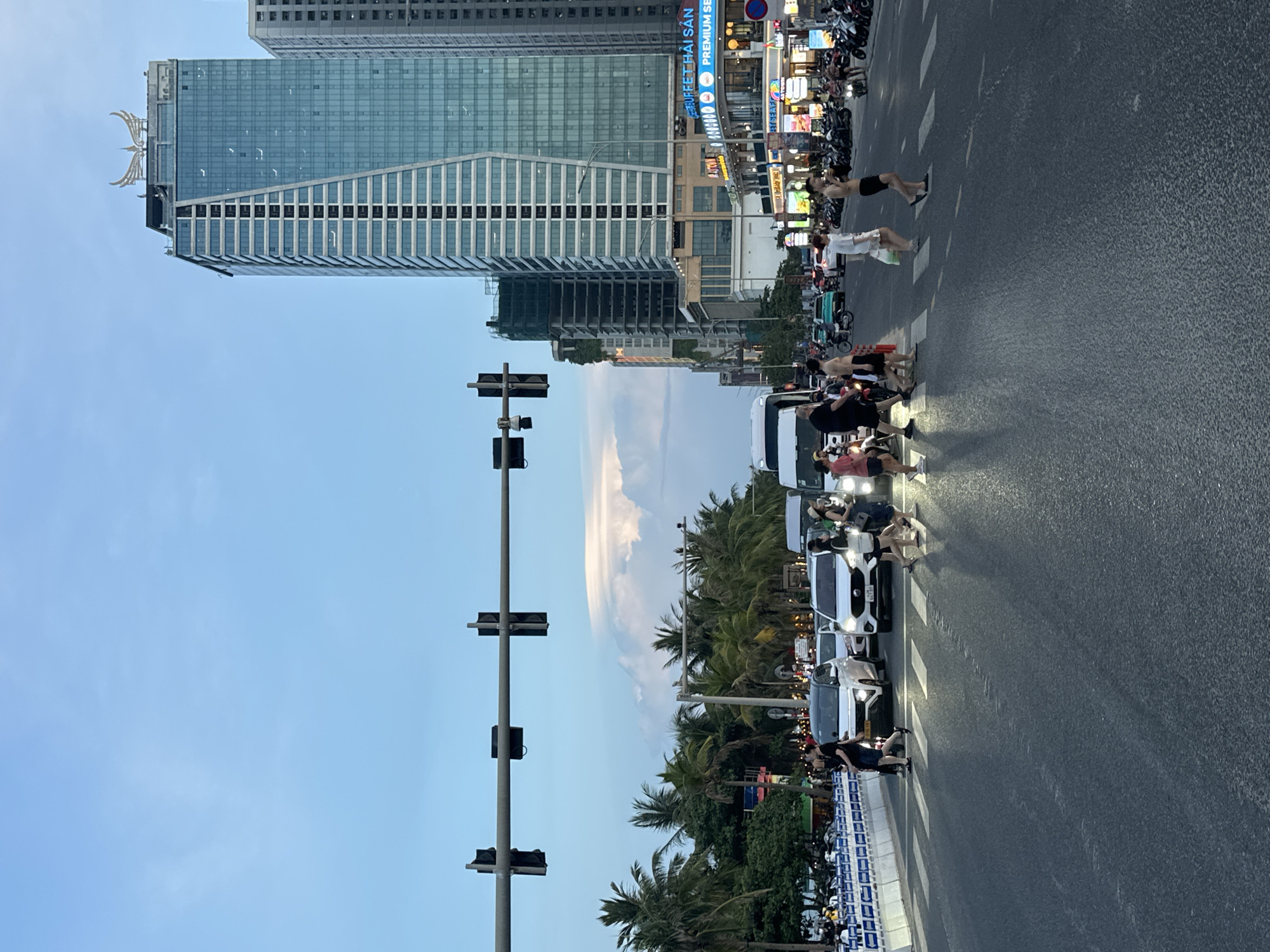 This is a vertical, eye-level photograph taken in Đà Nẵng, Vietnam, likely during the late afternoon or early evening given the lighting. The scene captures a bustling urban street with a prominent modern skyscraper on the left, featuring a distinctive metallic eagle sculpture at its apex. The skyscraper's glass facade reflects the sky and surrounding structures. Adjacent to the skyscraper, on the right side of the frame, is a strip of shops and businesses with bright, colorful signage, indicating a commercial area.
The foreground is dominated by a street with a pedestrian crossing. Several people are depicted crossing the street, some appearing to be walking, while others are standing or in motion, suggesting they are navigating the crossing. A line of vehicles, including cars and numerous motorcycles, are stopped or moving slowly, typical of city traffic. The vehicles have their headlights on, which contributes to the sense of dusk.
Palm trees and other lush greenery are visible behind the pedestrian area, suggesting a tropical or subtropical climate and proximity to a coastal or park-like environment. A traffic light pole with multiple signal heads stands in the middle ground, indicating a controlled intersection. The sky is a clear blue with some wispy white clouds.
The atmosphere appears lively and dynamic, with a mix of urban development, natural elements, and human activity. The presence of many motorcycles and the style of the buildings and storefronts are characteristic of Southeast Asian cityscapes. Some text is visible on the shop signs, but it is mostly illegible due to the image quality and distance.
