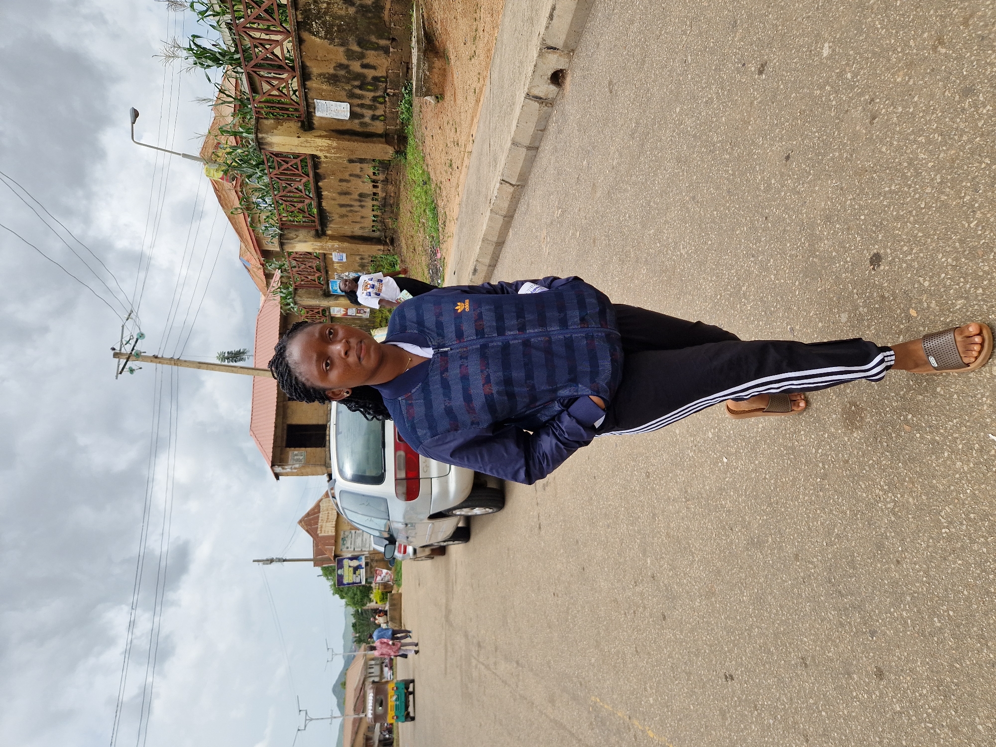 A young woman with braided hair walks on a paved road in Kabong, Nigeria. She is wearing a blue bomber jacket with horizontal stripes and black track pants with white stripes down the sides. She is also wearing brown sandals. The road is lined with a concrete sidewalk and a wall with a fence. In the background, there are buildings, vehicles, and power lines. The sky is cloudy.