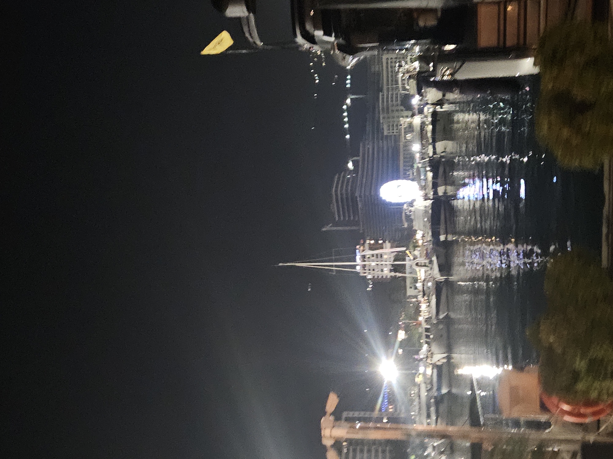 The image depicts a nighttime scene at a marina, likely in Budva, Montenegro, given the reference to the city. The water in the foreground is dark and reflects the lights from the surrounding area, creating a shimmering effect. Several boats, primarily yachts and sailboats, are docked in the marina. In the background, buildings and structures are visible, illuminated by various lights, suggesting a city skyline. A large Ferris wheel can be seen lit up in the distance. A yellow flag is prominently displayed on a structure on the right side of the frame. The overall atmosphere is that of a bustling, illuminated harbor at night. There are no visible people in the frame.
